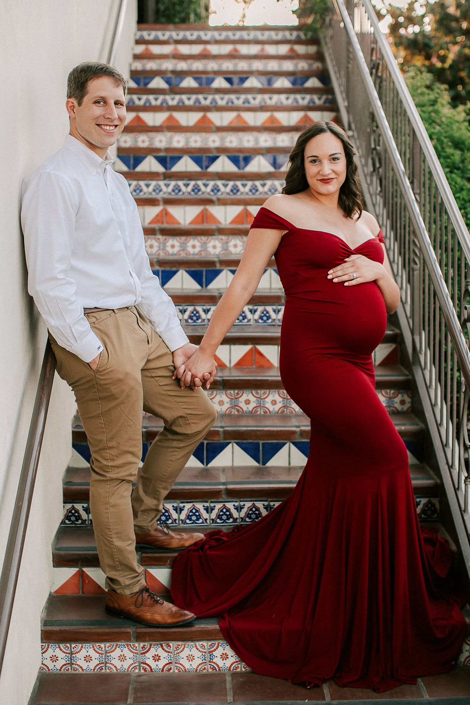 Pregnant couple poses on colorful tiled stairs, woman in red maternity gown holding partner's hand.