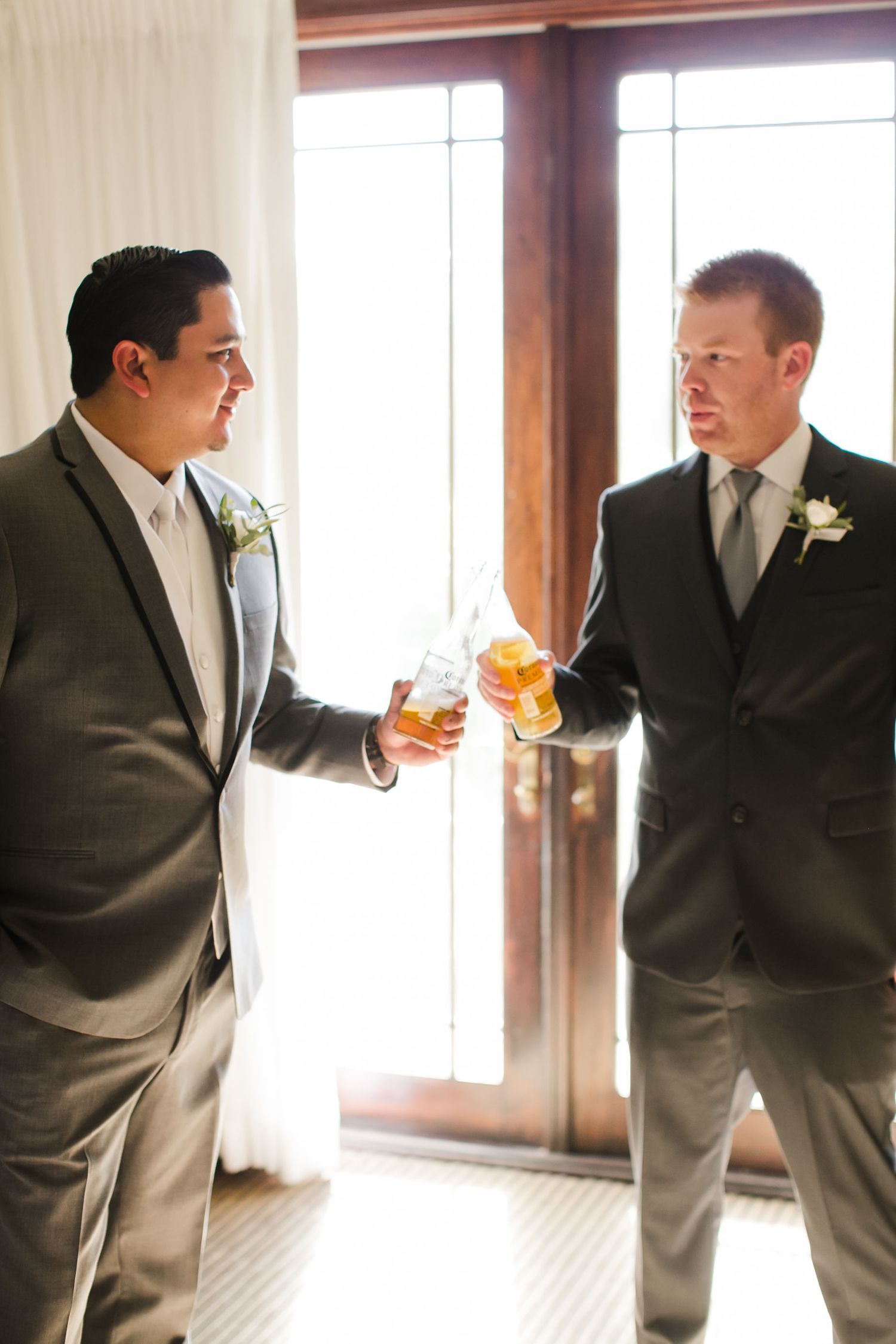 Two groomsmen in gray suits toasting with whiskey glasses near sunlit doors adorned with white boutonnieres at a wedding.