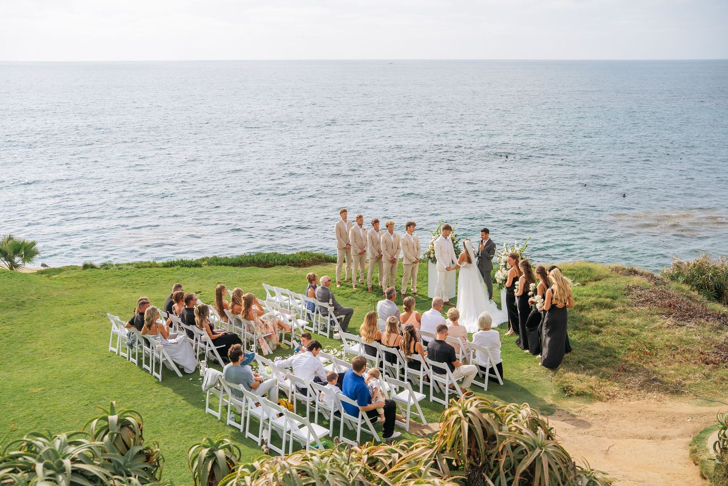 Outdoor oceanfront wedding ceremony with guests seated in white chairs, bridesmaids, and groomsmen overlooking the sea.