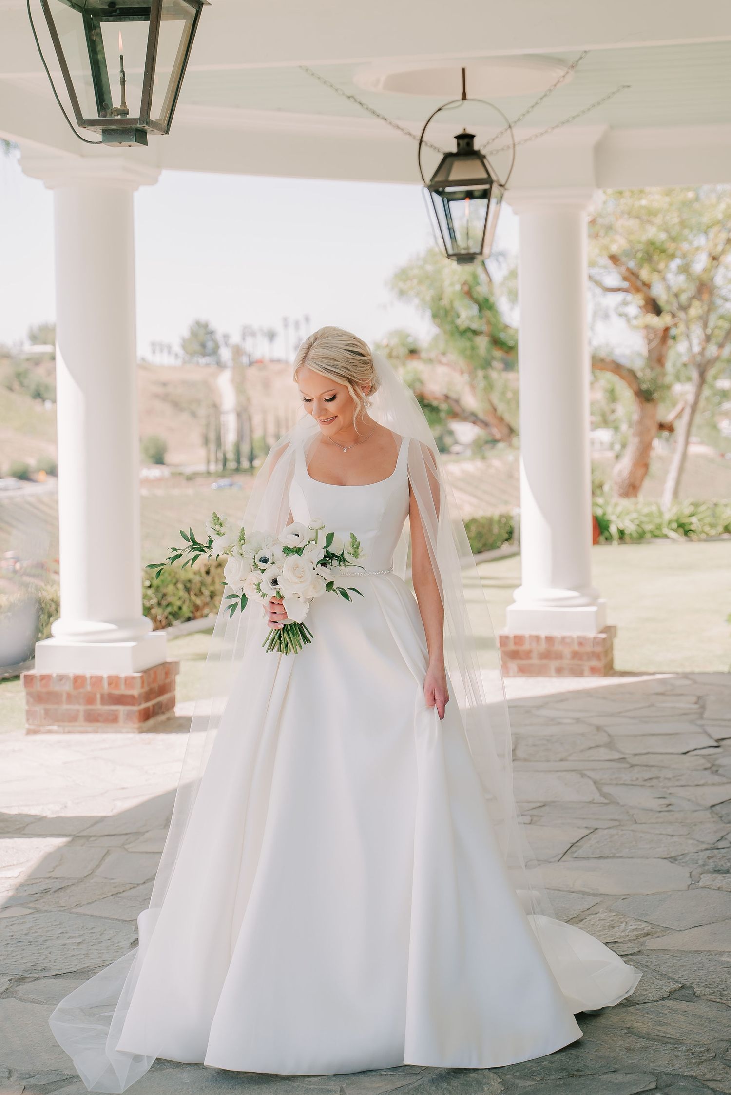 Elegant bride in classic white ball gown with veil holding floral bouquet under columned portico outdoors.