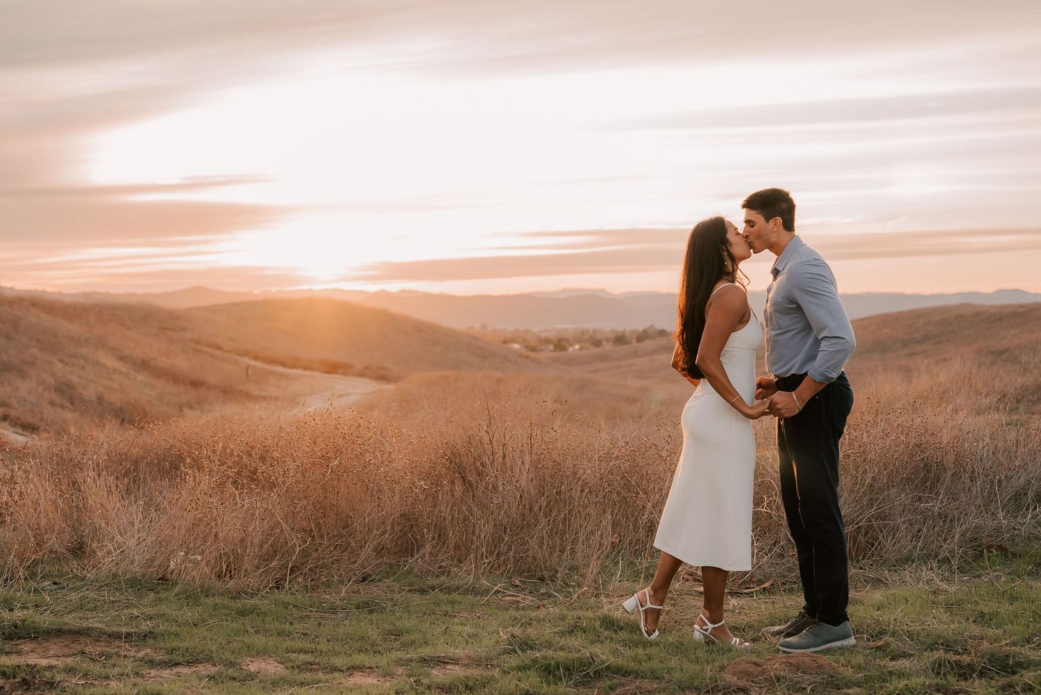 Couple in a white dress and gray button-up shirt share a kiss on a grassy hillside at golden hour sunset.