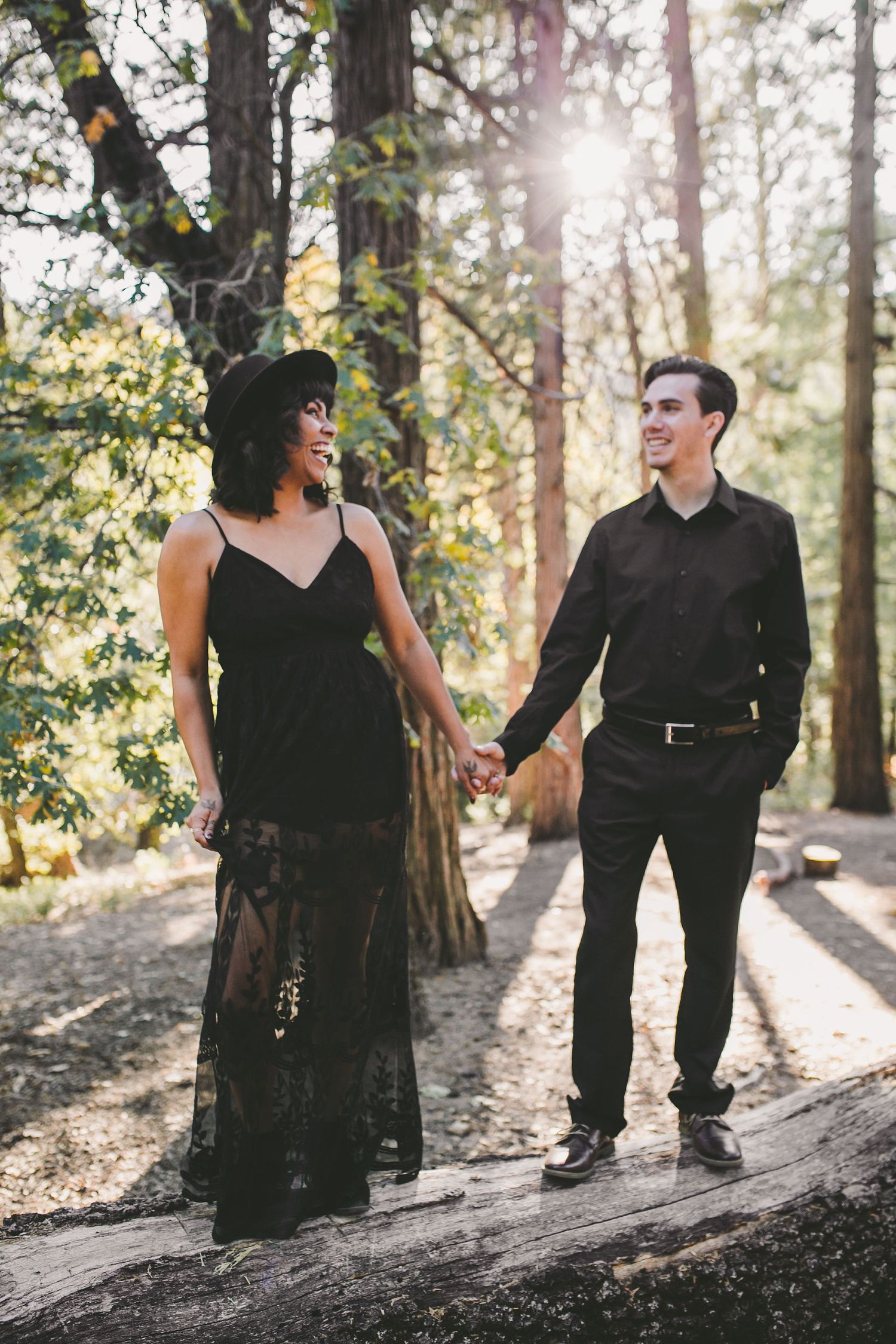 Couple in matching black outfits holding hands in a sunlit forest, woman in lace gown and hat, man in dress shirt.