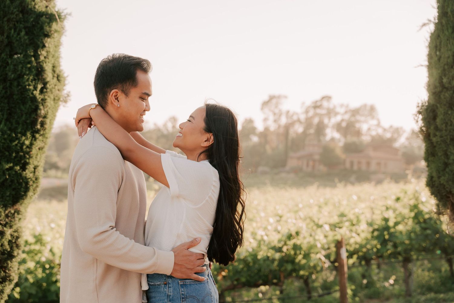 Couple embracing romantically in a sunlit vineyard surrounded by lush greenery and cypress trees.