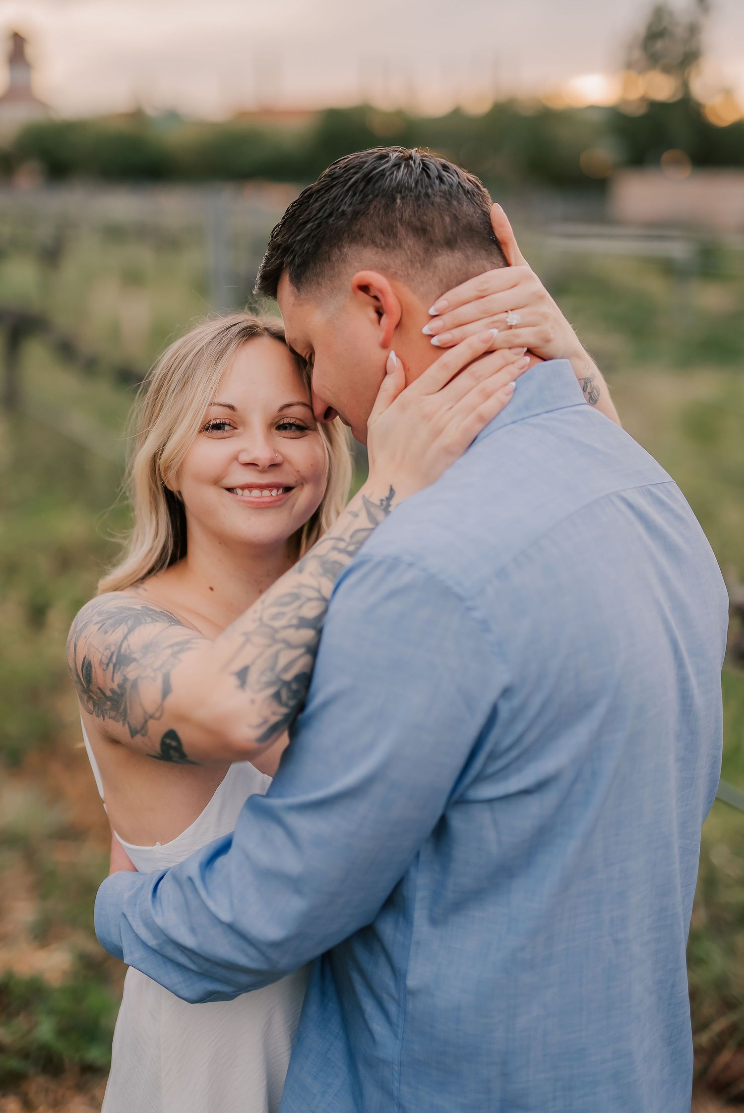 A smiling blonde woman with tattoos embraces a man in a blue shirt outdoors at sunset during an engagement photo session.
