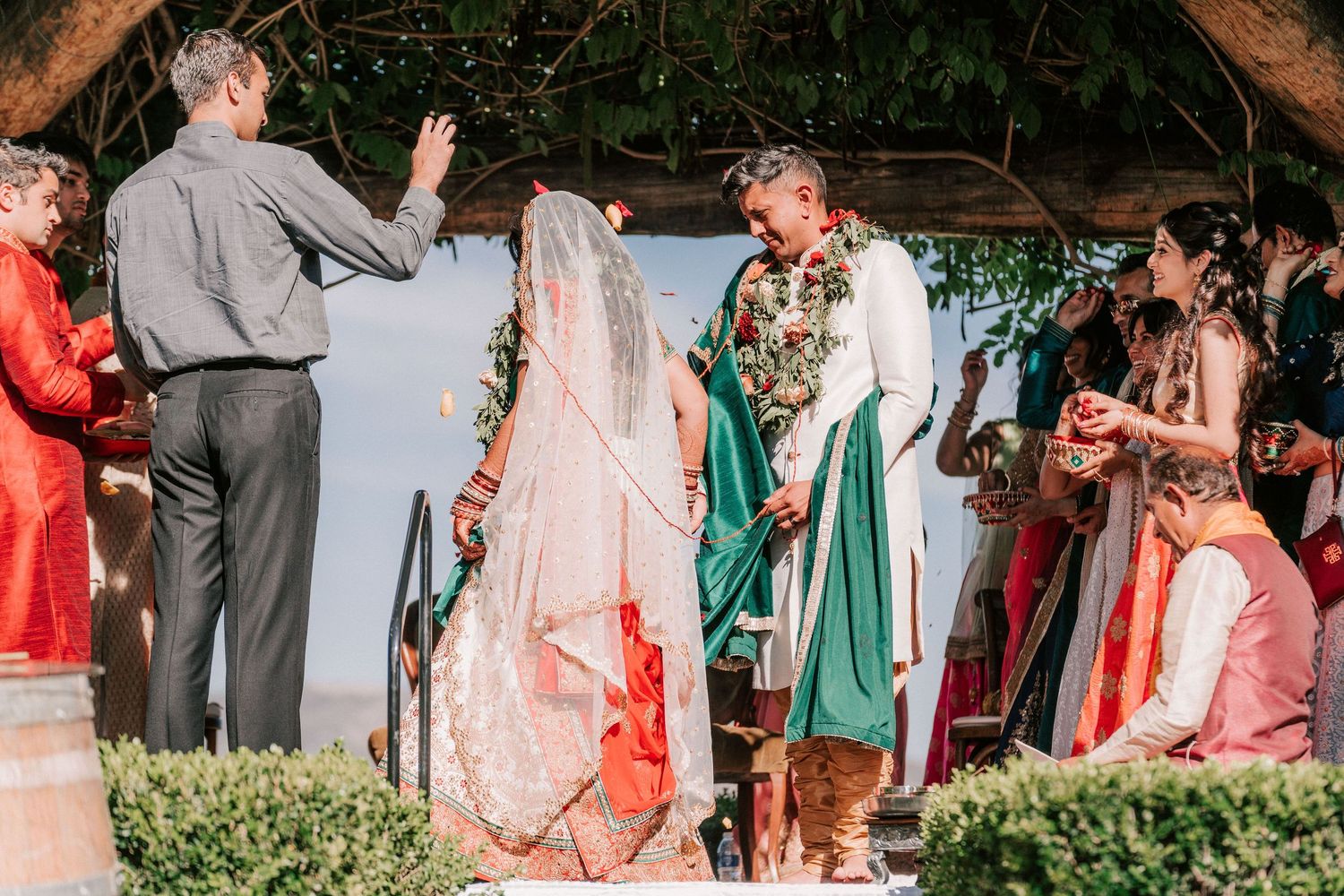 Indian bride and groom exchanging vows under pergola with officiant and guests.