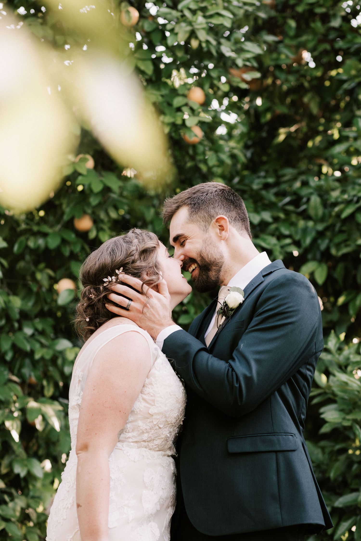 A smiling groom in a navy suit tenderly holds his bride's face as they share a romantic moment outdoors near lush greenery.
