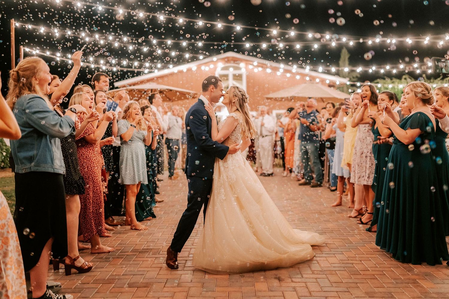 Bride and groom share a kiss while guests celebrate with confetti and bubbles at an outdoor evening wedding reception.