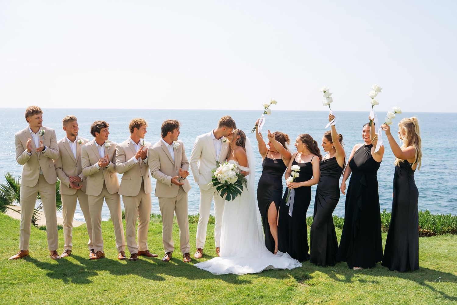 Wedding party poses oceanside, groomsmen in tan suits and bridesmaids in black gowns cheering as couple kisses.