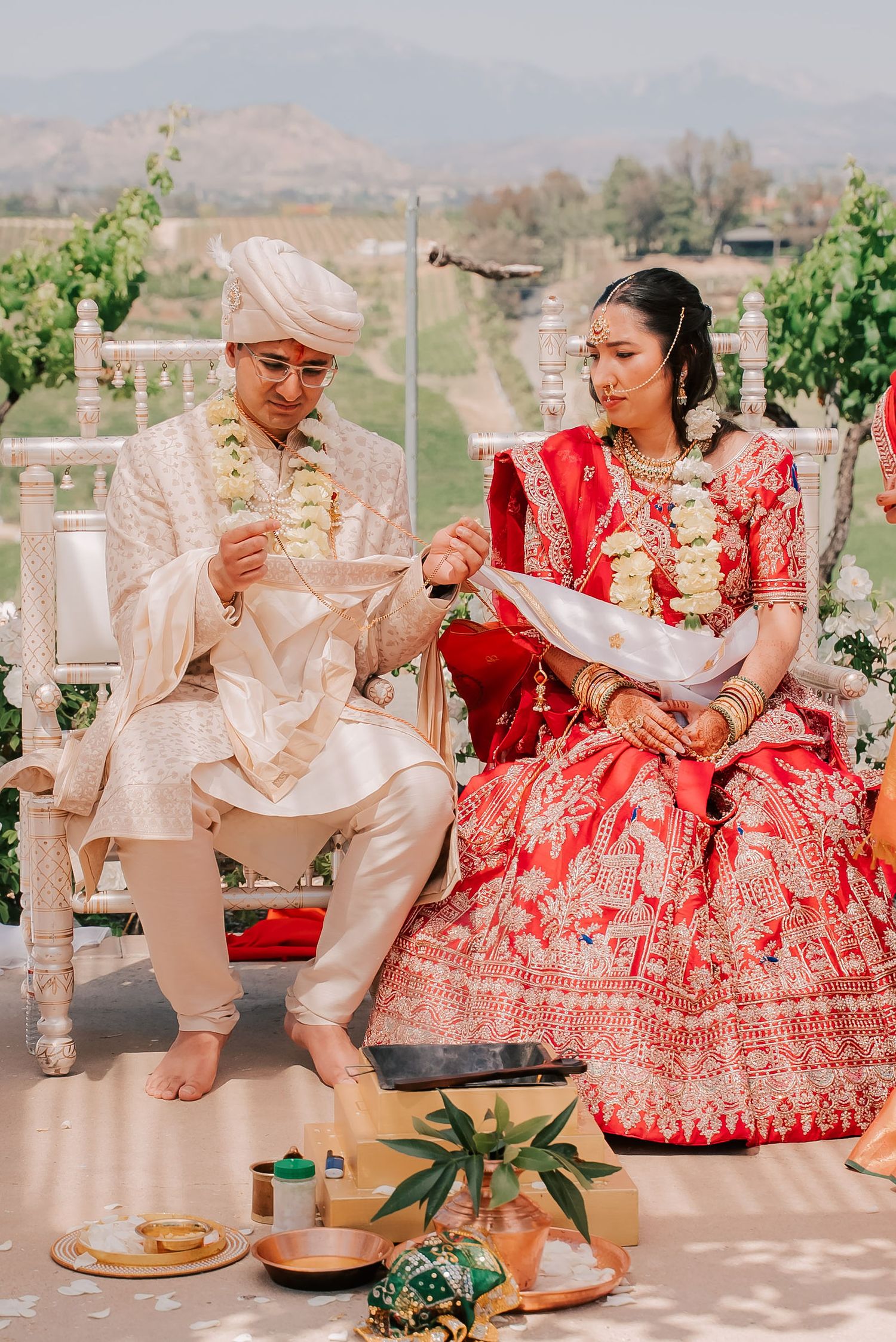 Indian bride and groom in red and cream attire performing traditional Hindu wedding rituals outdoors.