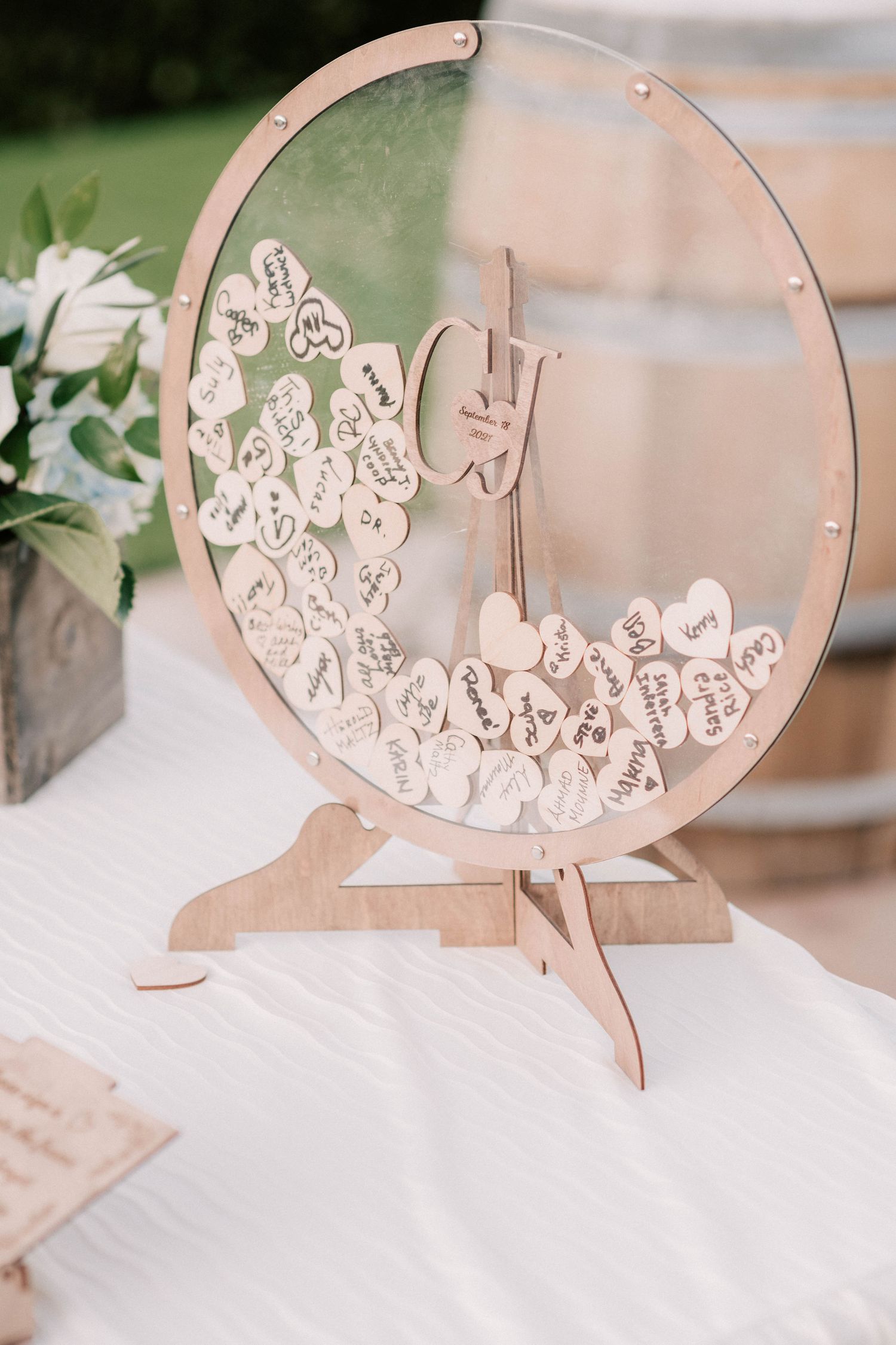 Wooden circular wedding guest book drop box with heart-shaped cards on a white table at an outdoor reception.