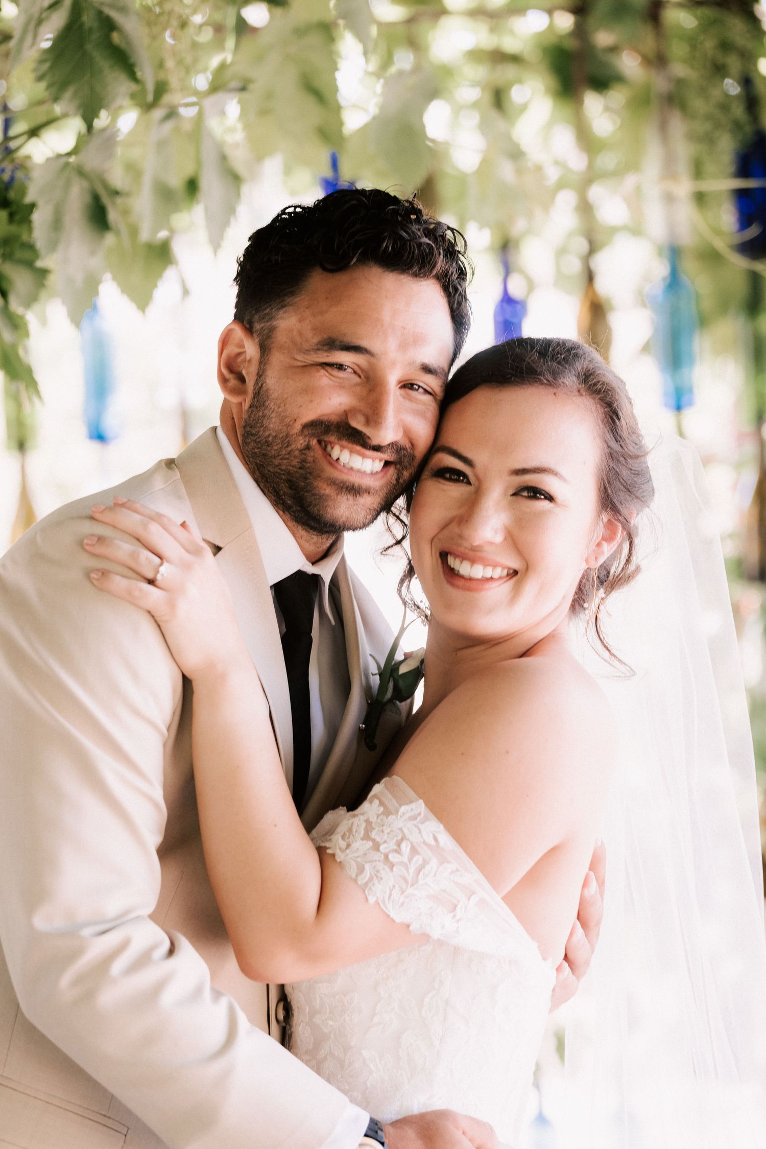 A smiling bride and groom embrace outdoors under lush greenery and blue glass decorations on their wedding day.