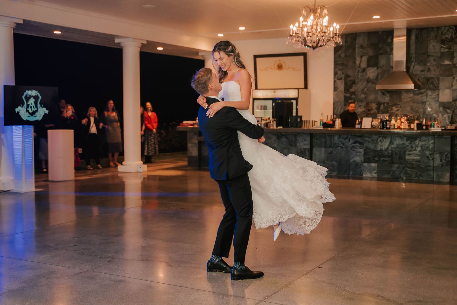 Groom lifts bride in wedding dress during romantic first dance in elegant ballroom with chandelier.