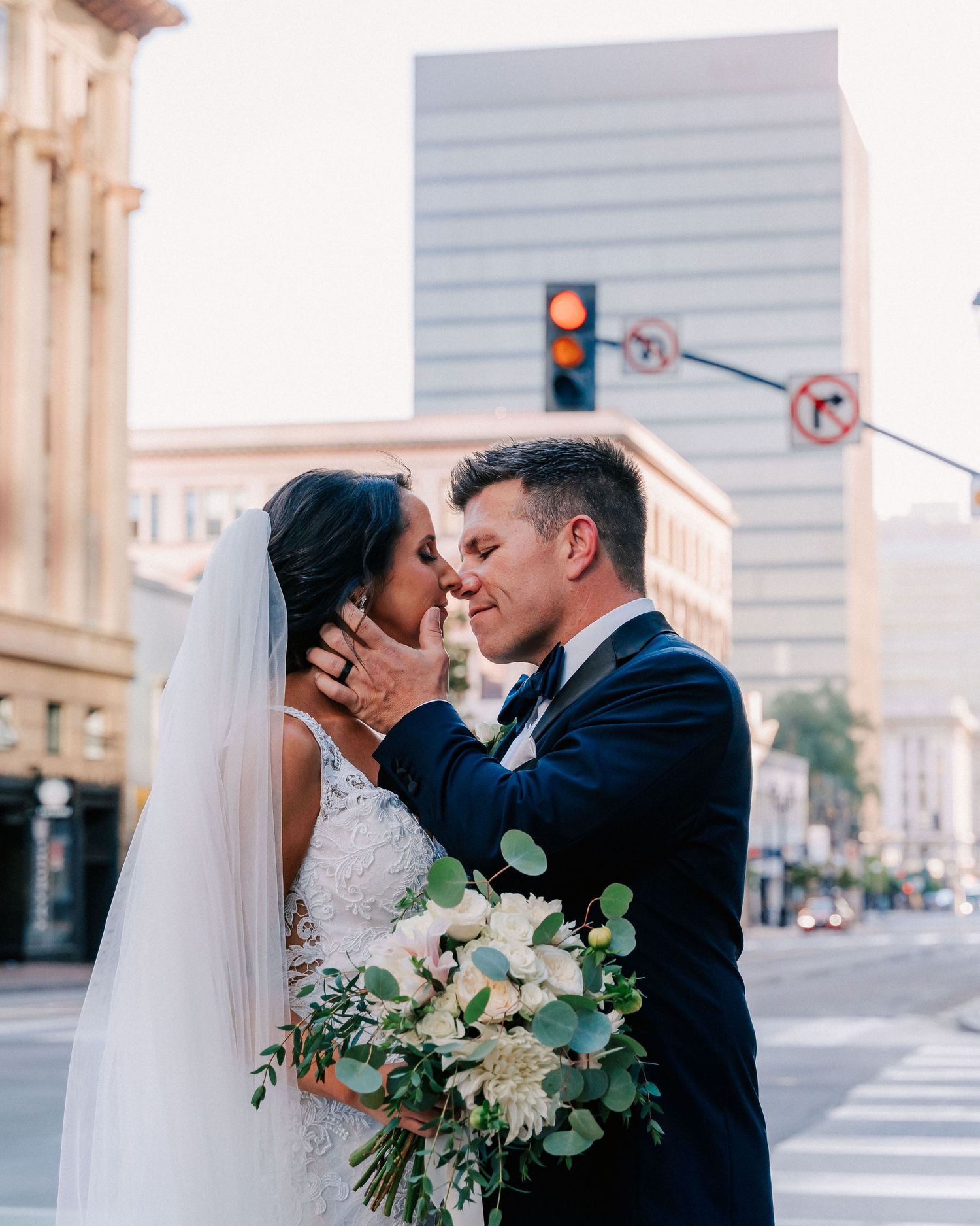 A bride and groom share a romantic kiss on a city street corner, with the bride holding a white floral bouquet.