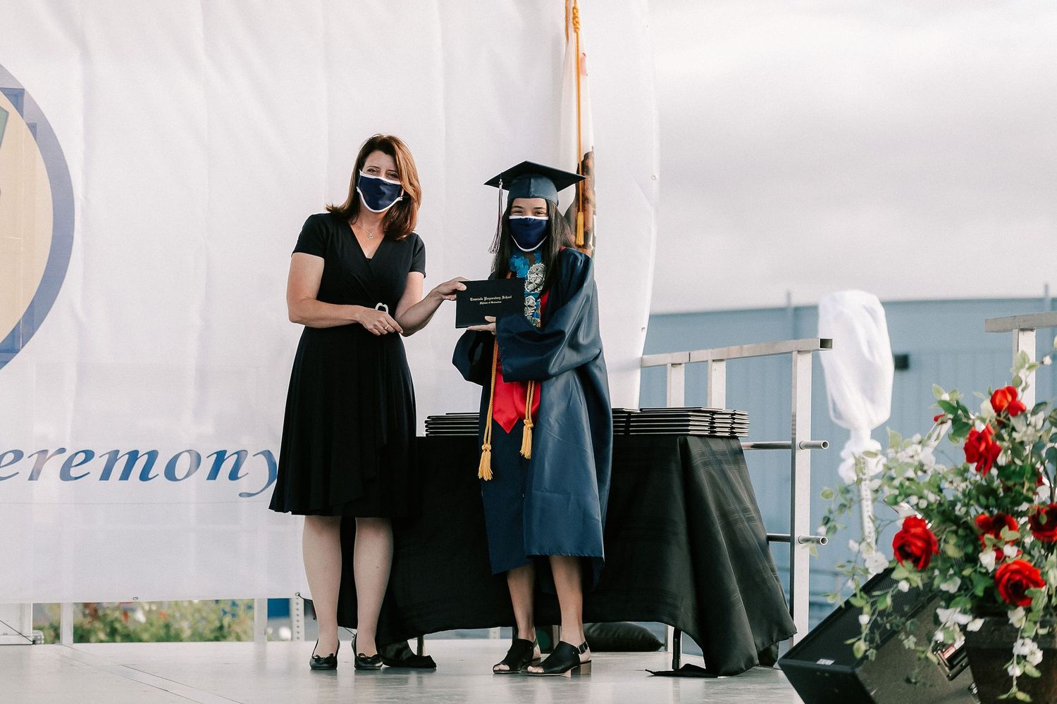 Graduate in cap and gown receives diploma on stage during outdoor graduation ceremony with masked presenter.