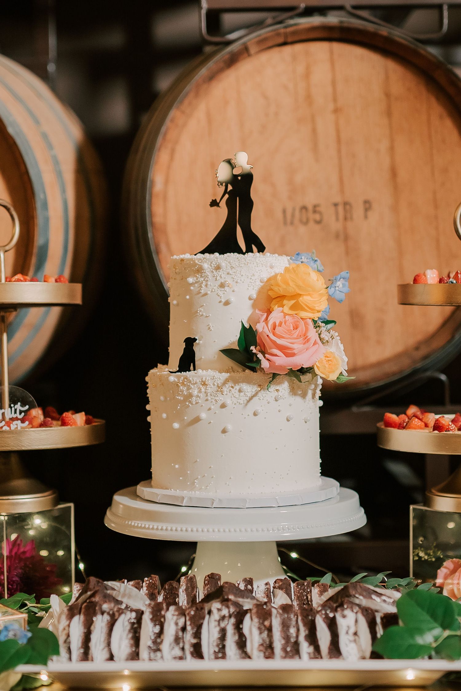 Two-tier white wedding cake with floral decor and couple topper, displayed near wine barrels at a winery reception.