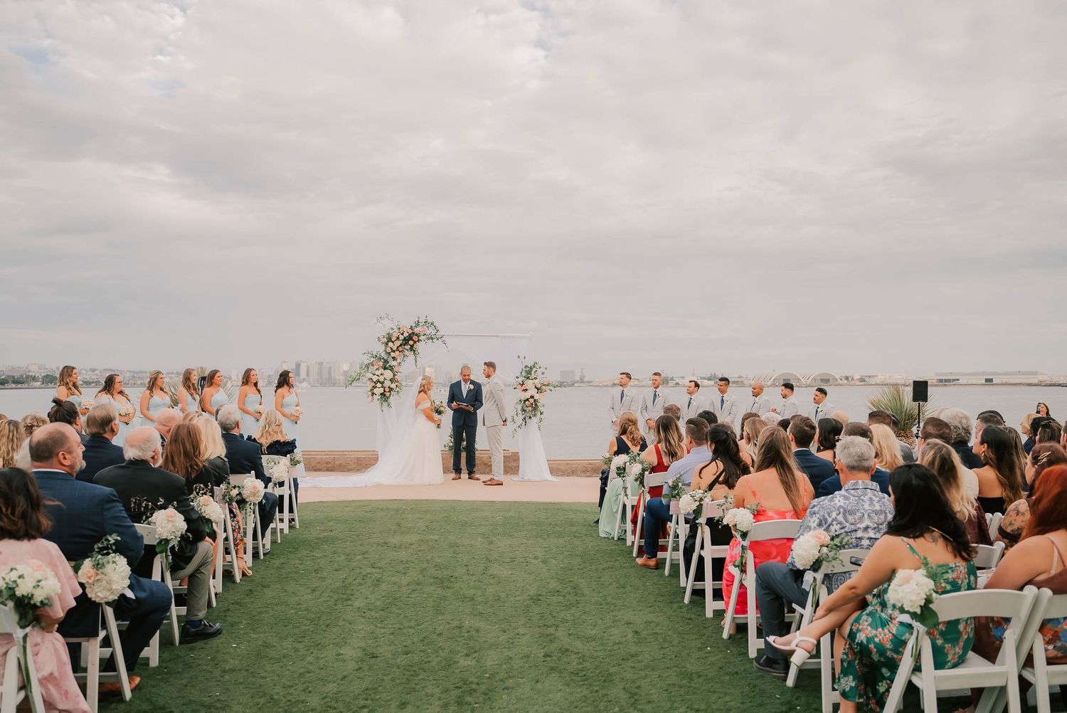Wedding guests seated on white chairs watch a couple exchange vows at an outdoor waterfront ceremony with floral arch and bridal party.