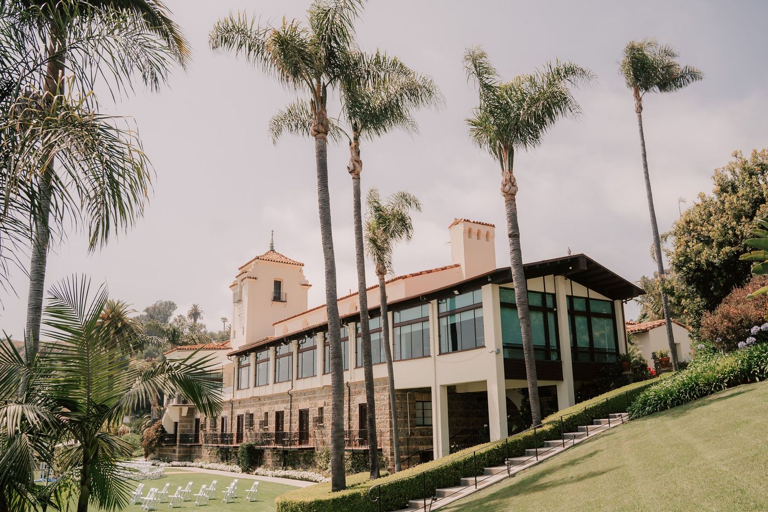 Elegant Mediterranean-style building with a tower, large windows and terrace, surrounded by tall palm trees and manicured green lawns.