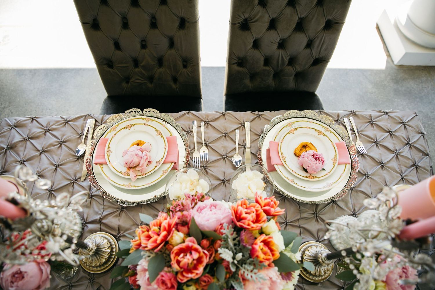 Elegant wedding table setting with pink floral plates, gold accents, and a vibrant peony centerpiece on a quilted tablecloth.