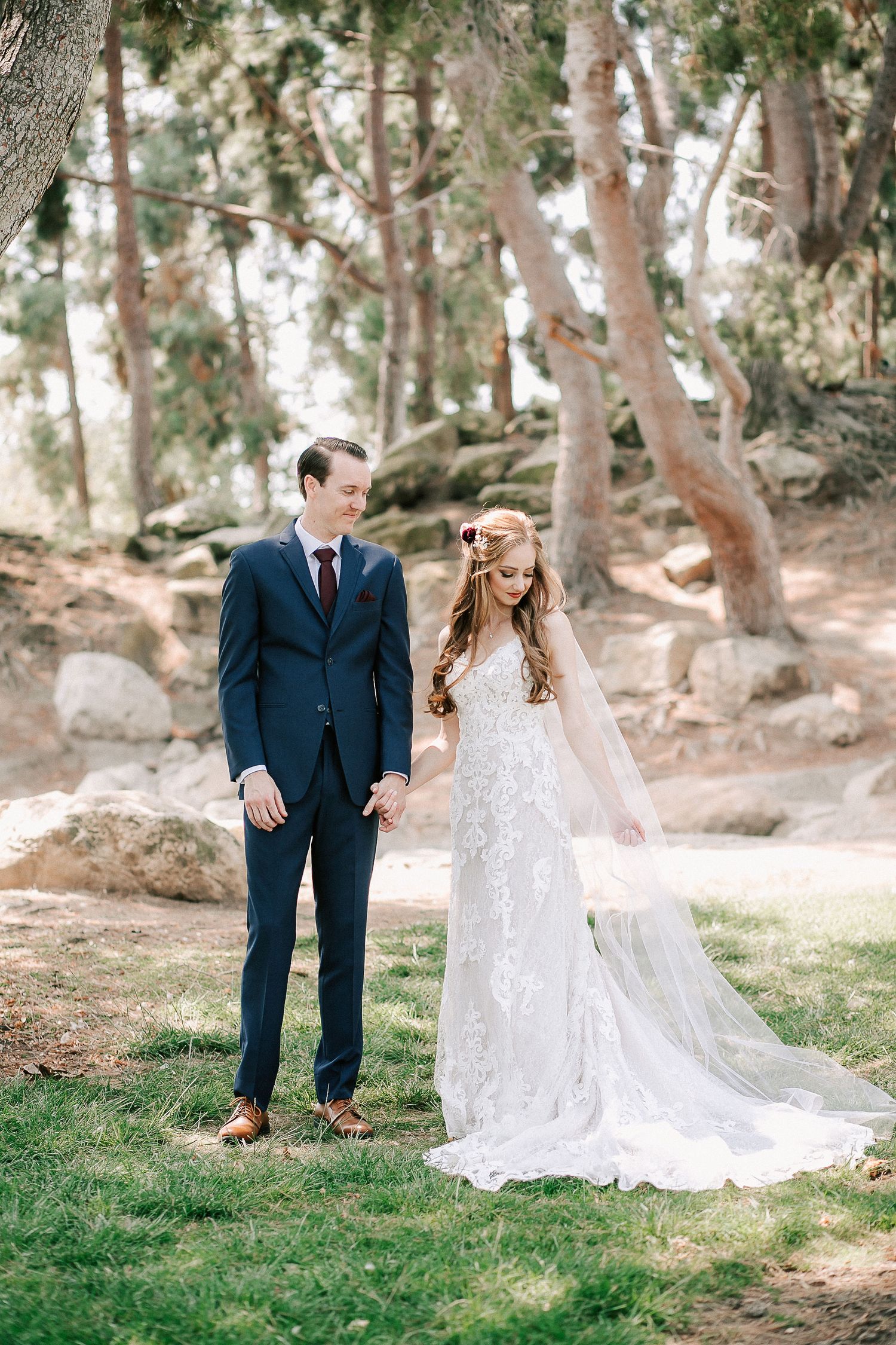 Bride in lace gown and groom in navy suit hold hands outdoors among trees on their wedding day.