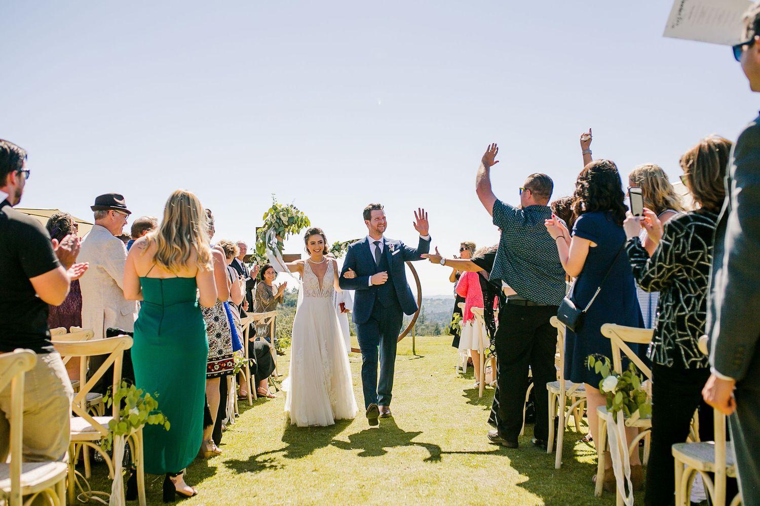 Newlywed couple walks down the aisle at an outdoor wedding ceremony as guests cheer under a clear blue sky.
