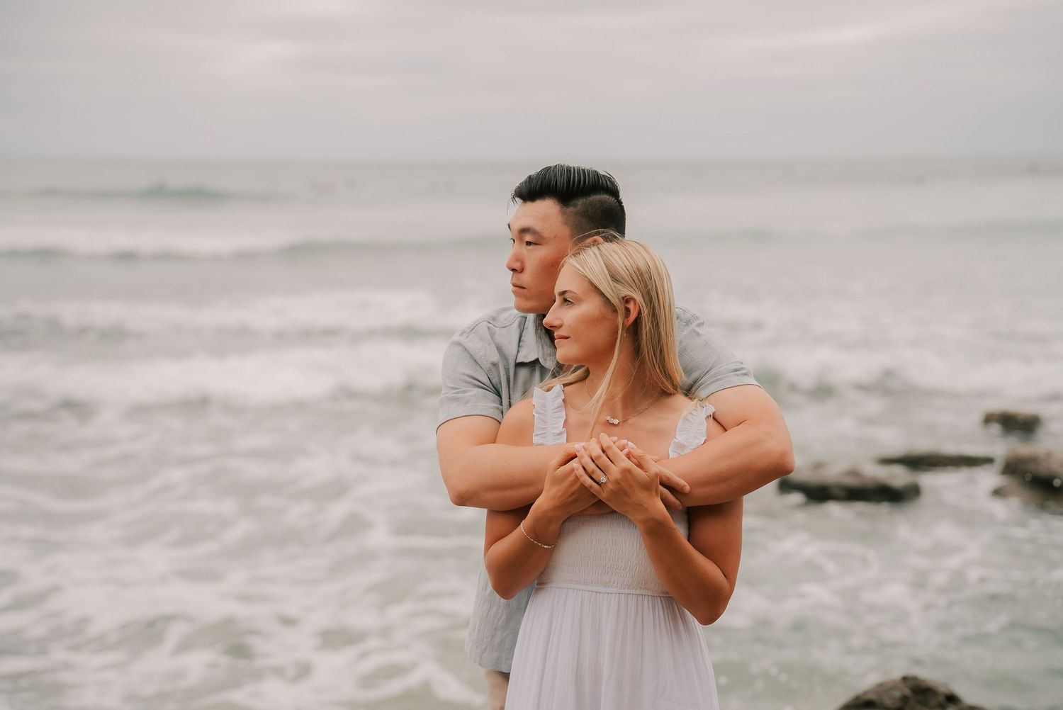 Couple embracing on a rocky beach, man holding woman from behind near ocean waves in a romantic engagement photo.