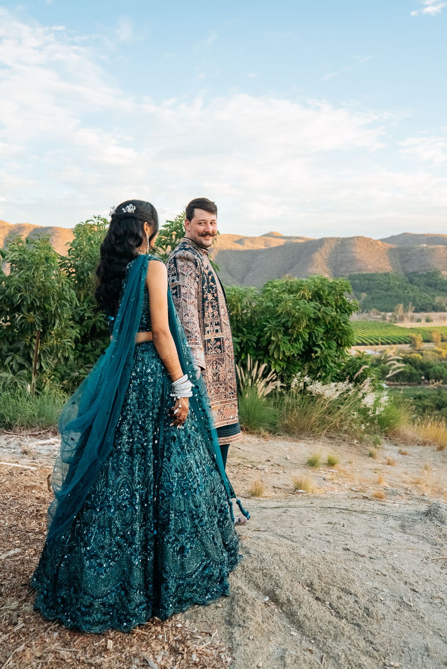 Indian bride in teal lehenga and groom in traditional attire pose outdoors with mountain vineyard backdrop.
