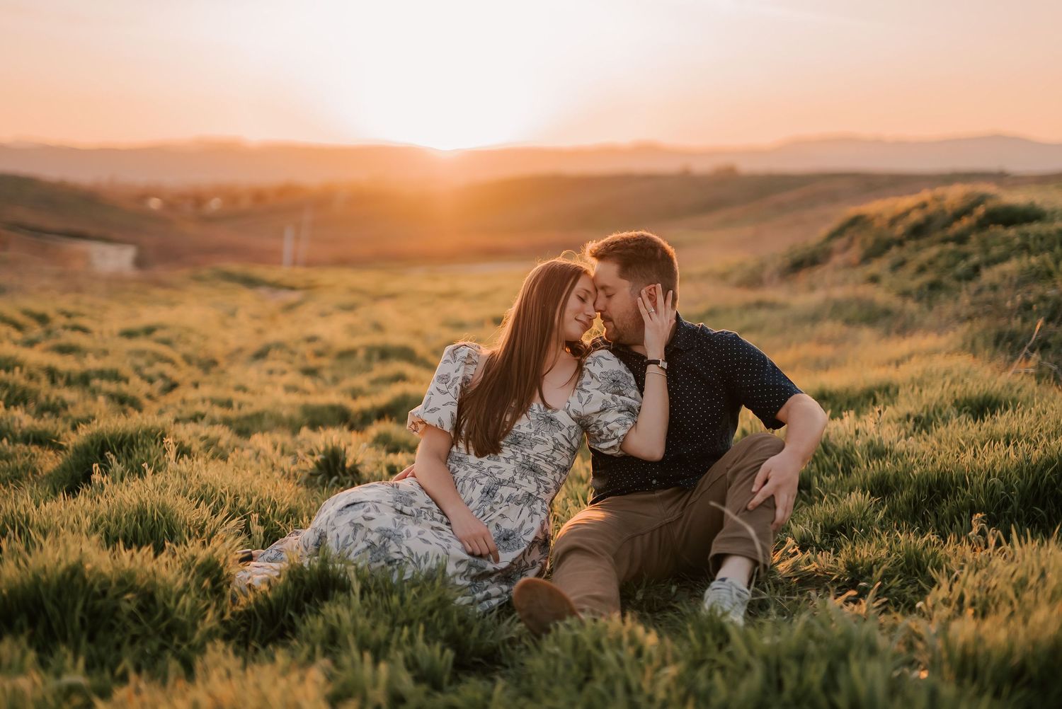 Couple sitting in green field sharing a kiss at golden sunset with rolling hills in the background.
