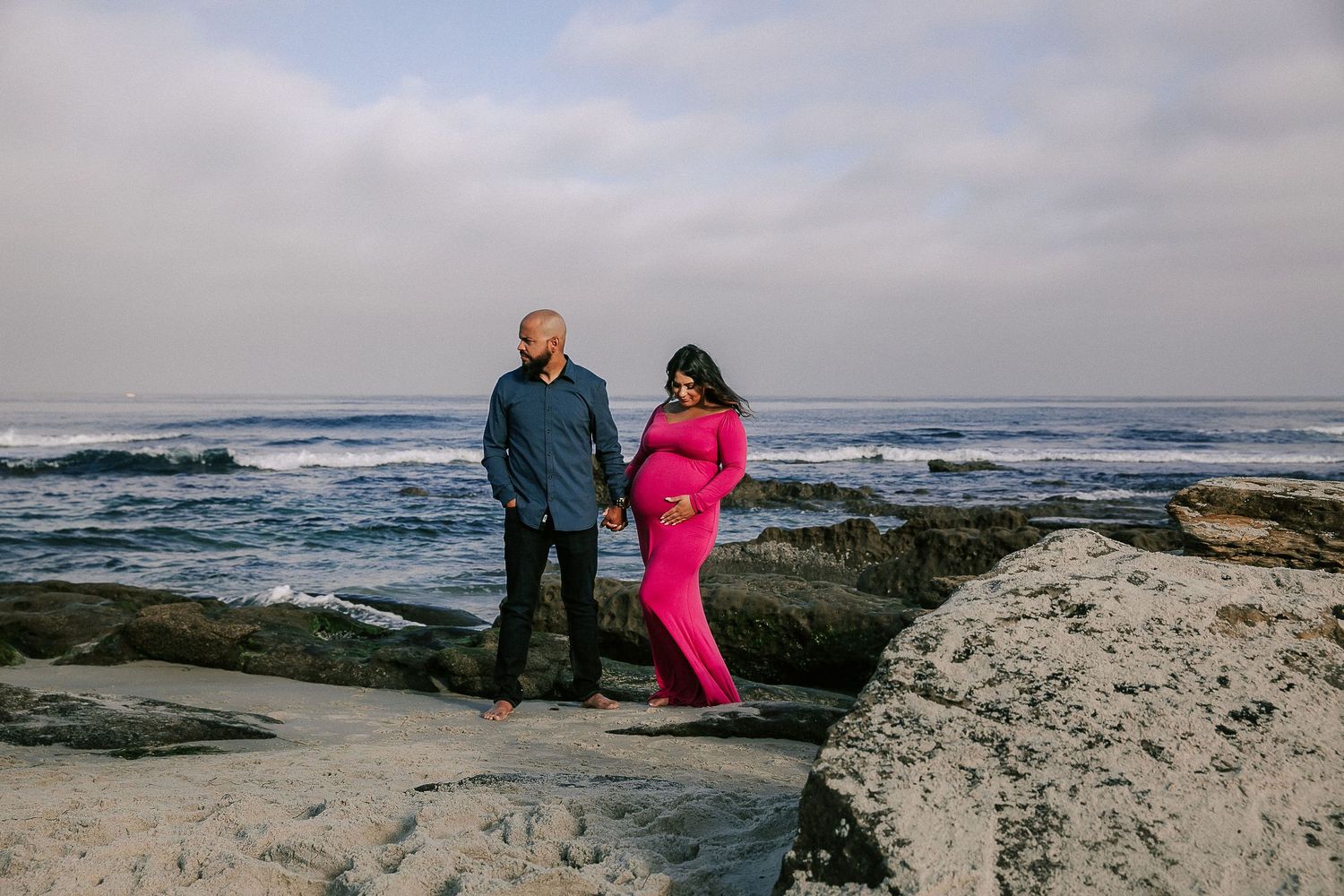 Couple holding hands on rocky beach, pregnant woman in pink gown, ocean waves behind them.