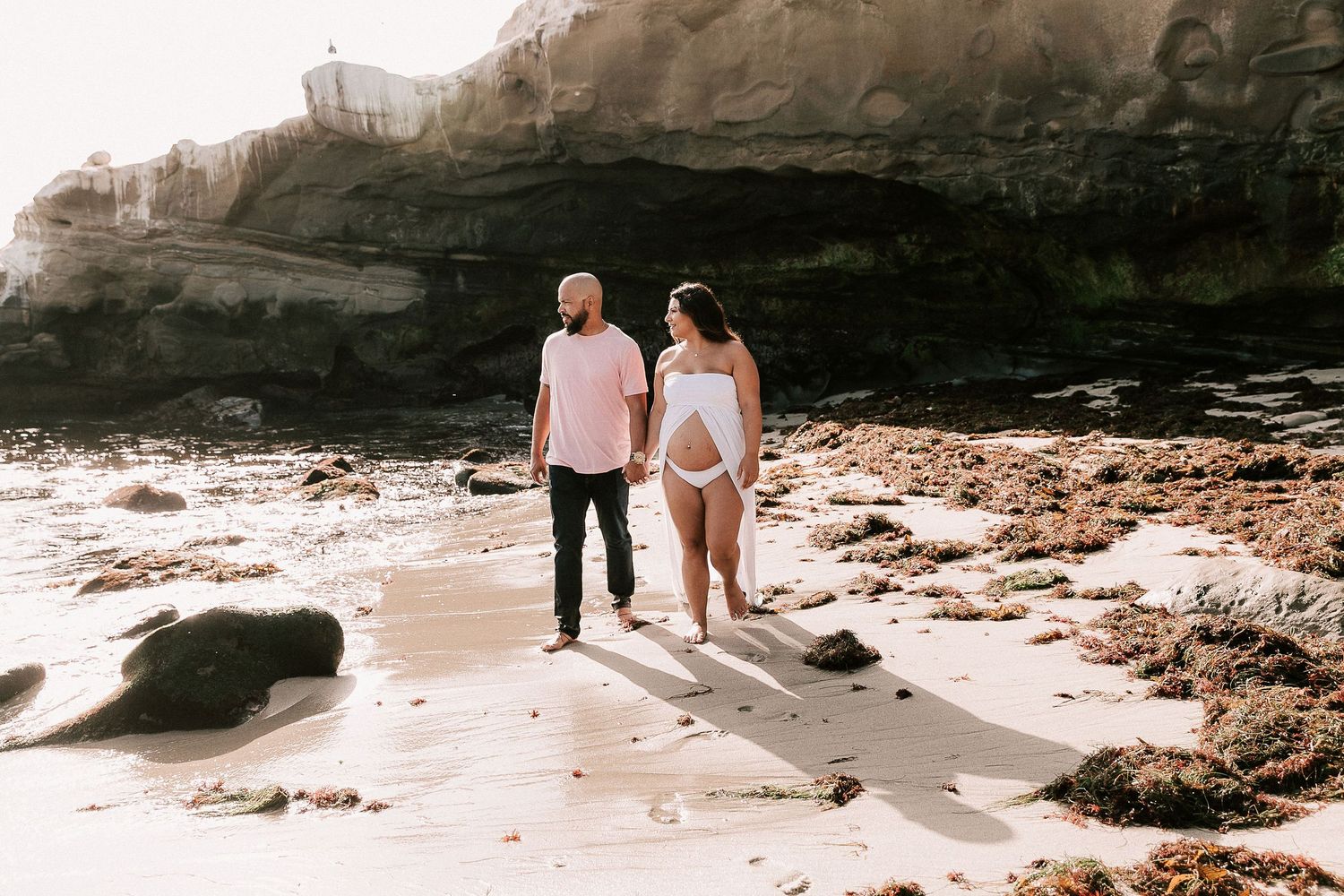 Expecting couple holding hands walking on sandy beach surrounded by rocks in warm golden light.