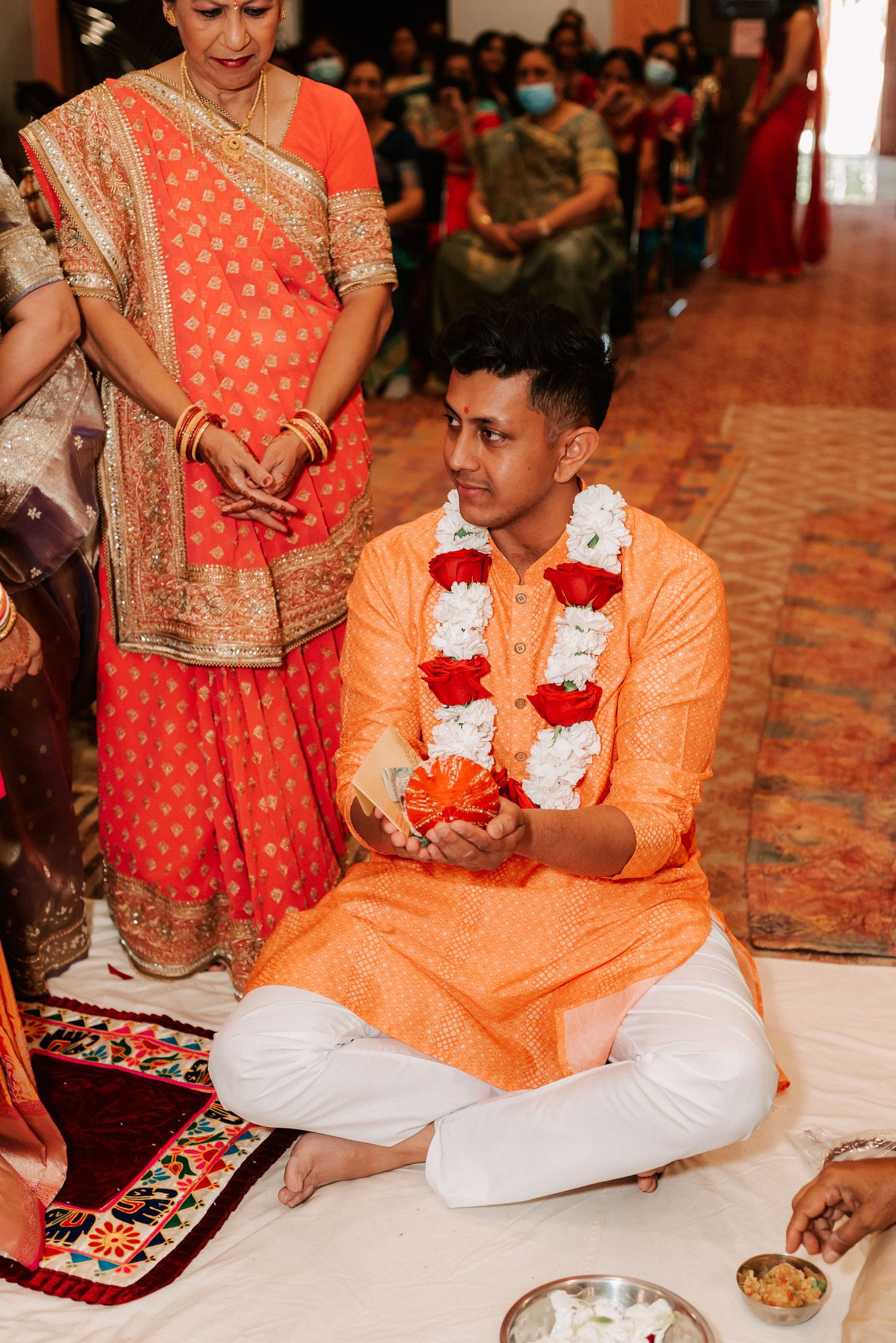 A groom in an orange kurta wearing a floral garland sits cross-legged during a Hindu wedding ceremony.