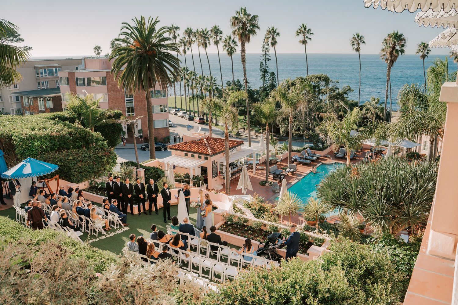 Aerial view of an outdoor wedding ceremony by a pool with ocean views, palm trees, and guests seated on white chairs.