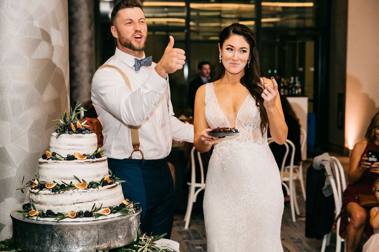 Bride and groom smiling beside a four-tier wedding cake decorated with greenery and flowers at their reception.