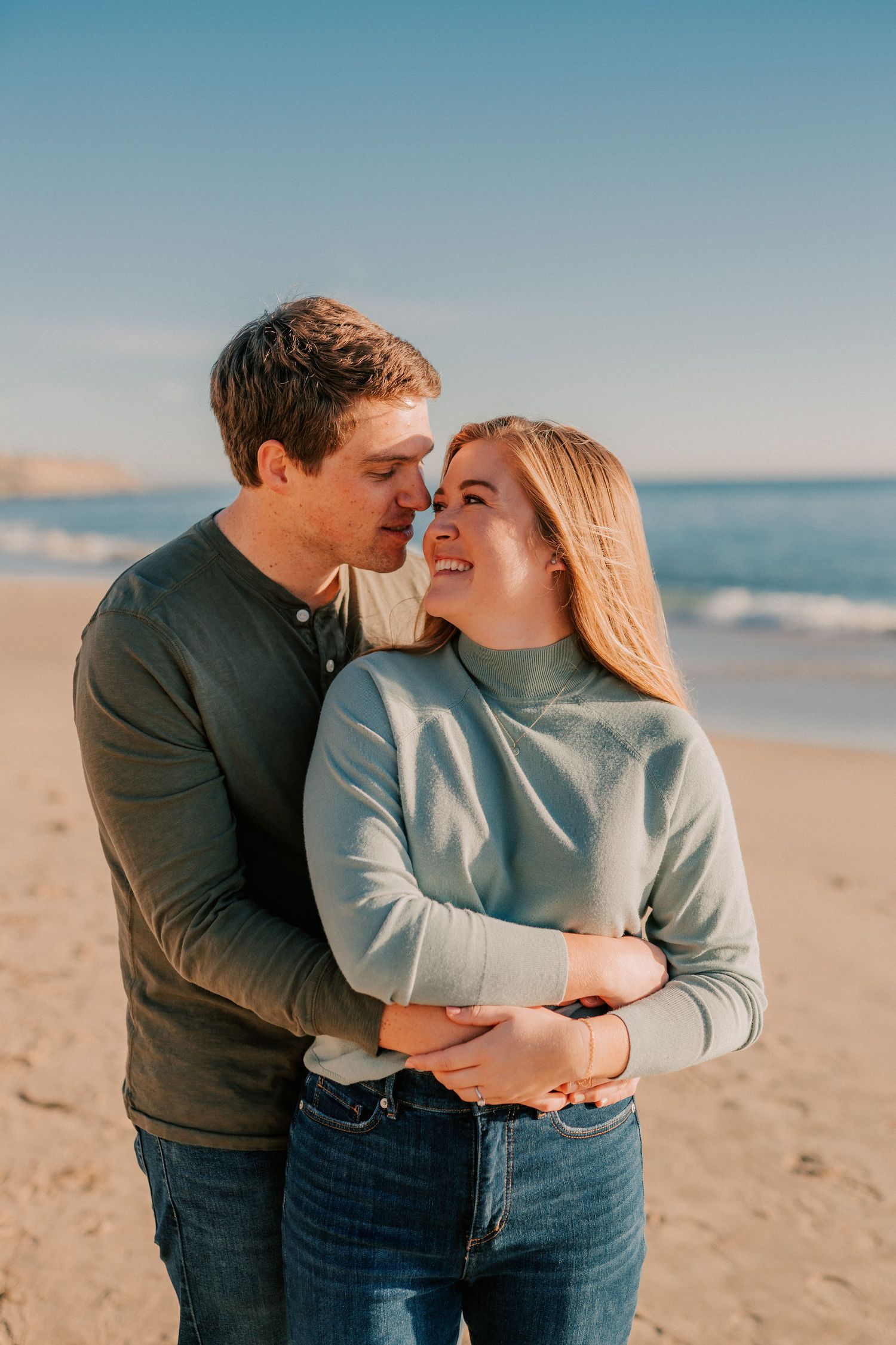 Happy couple embracing on sunny beach with blue sky and ocean waves behind them.