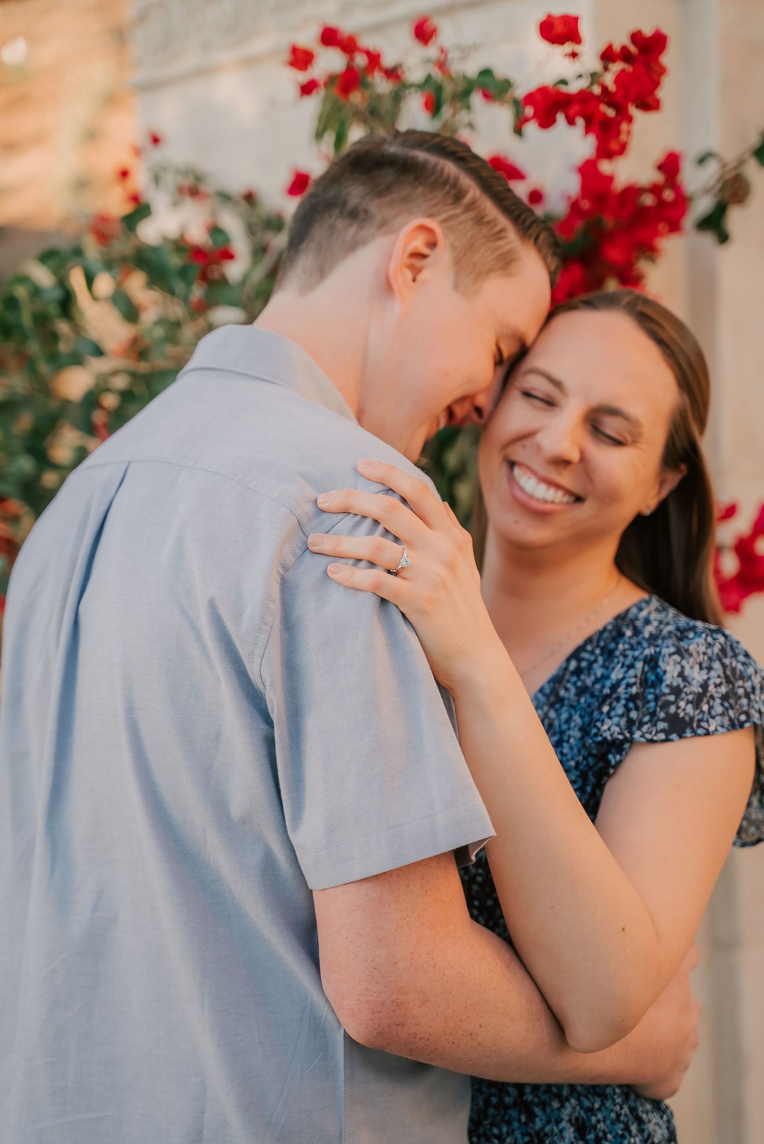A smiling couple embraces closely in front of red bougainvillea flowers, her eyes closed in joy as he leans his forehead against hers.