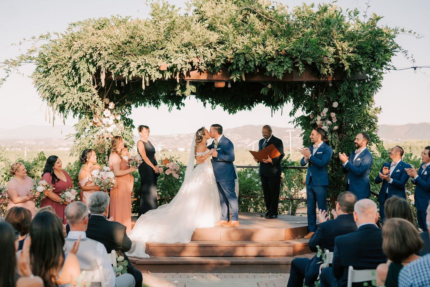 Couple shares first kiss at outdoor wedding ceremony under lush floral arbor with scenic vineyard views.