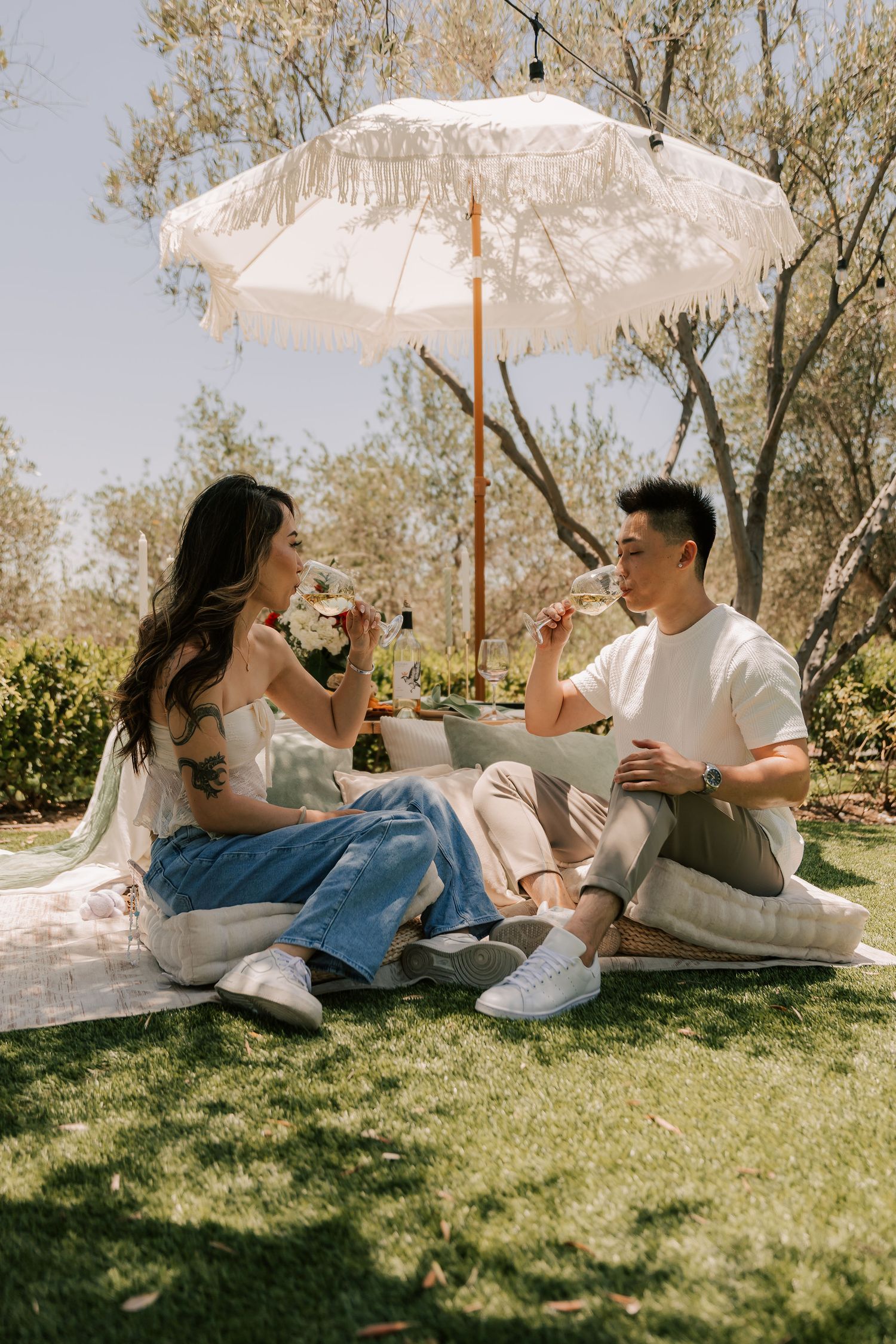 Couple enjoying wine at an outdoor picnic under a white fringed umbrella on a sunny day in a lush garden.