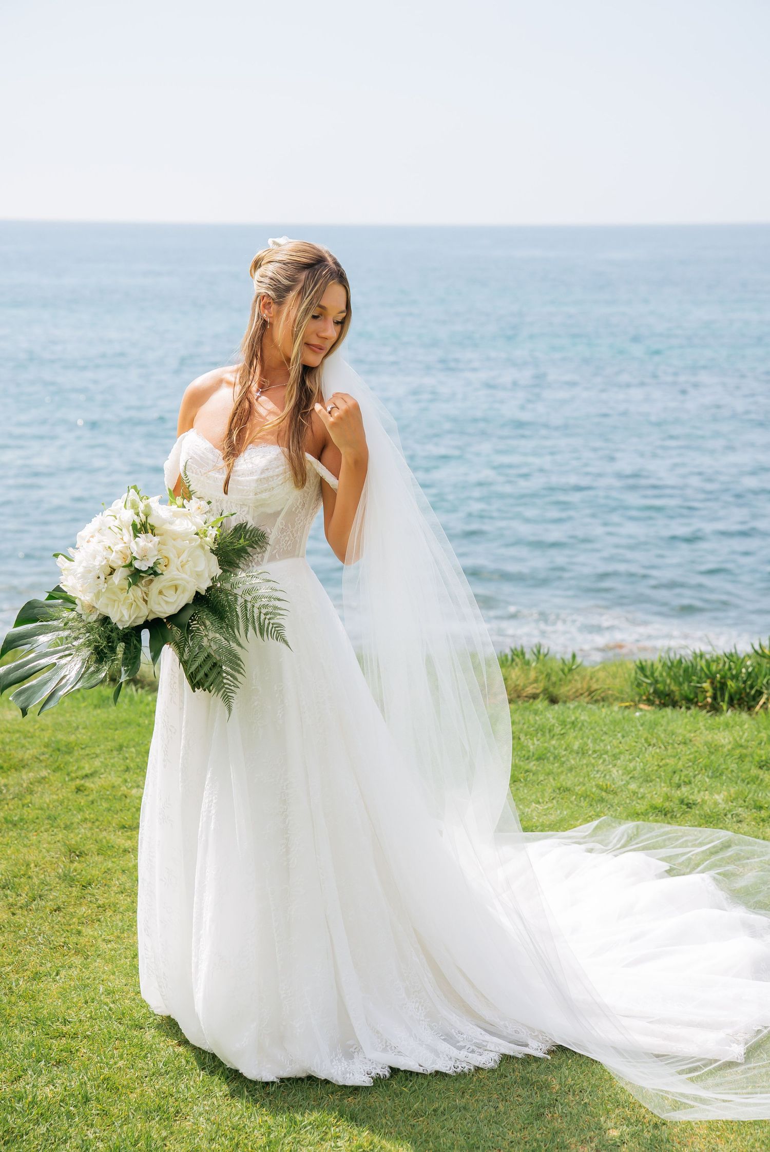 Elegant bride in a strapless white tulle gown holding a white floral bouquet with tropical leaves by the ocean.