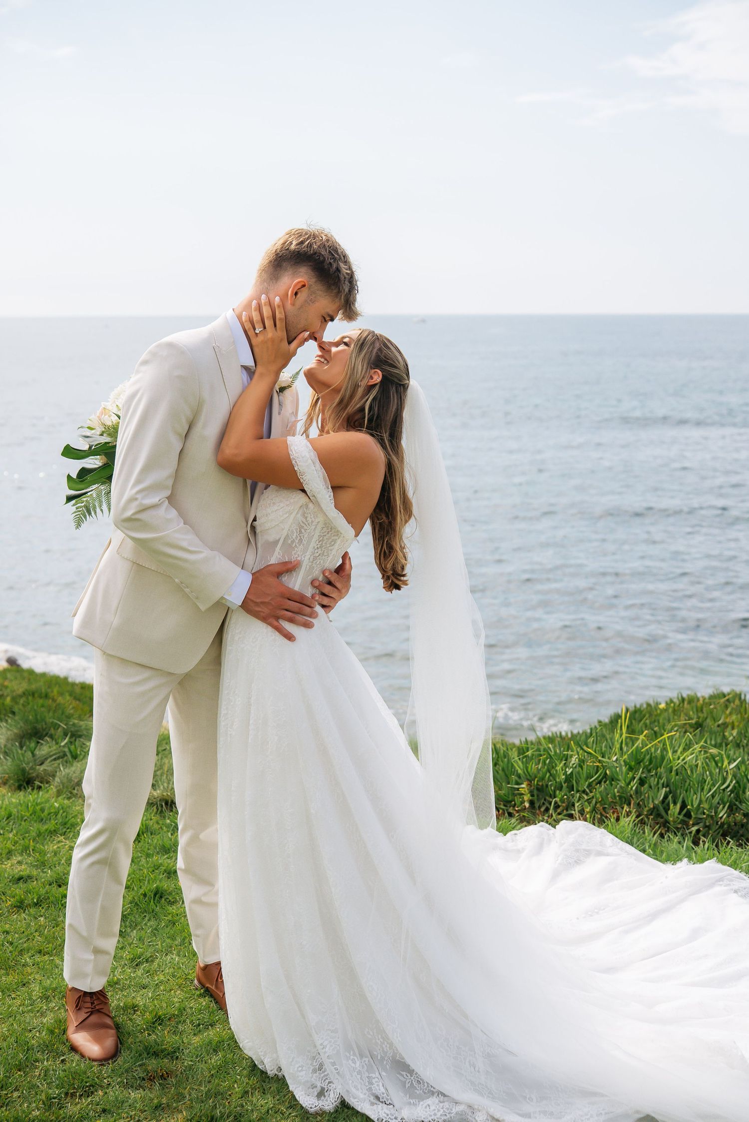 Bride and groom share a romantic kiss on a coastal cliff, with ocean views behind them on their wedding day.