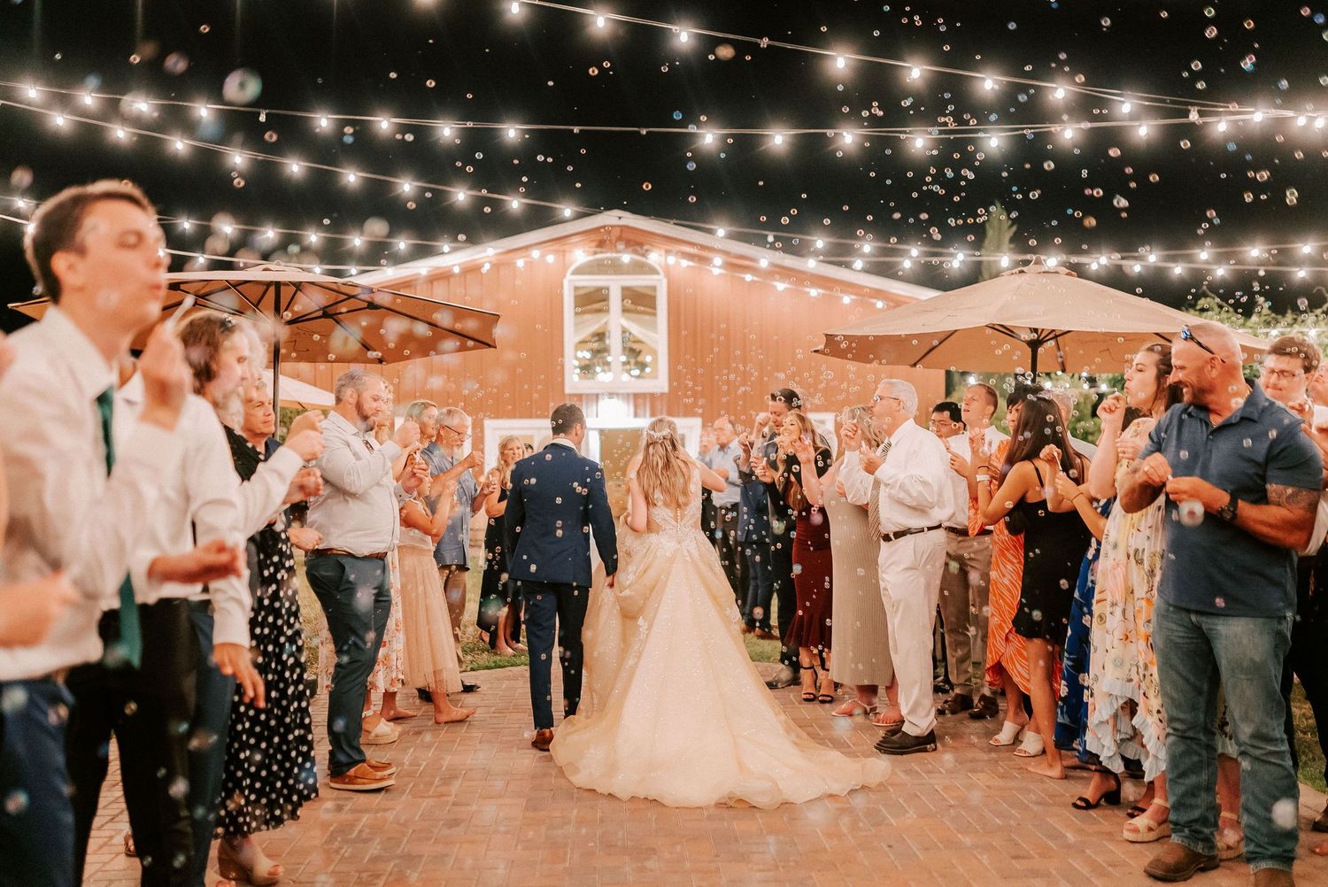Bride and groom walk through confetti celebration with guests cheering under string lights at outdoor barn wedding reception.
