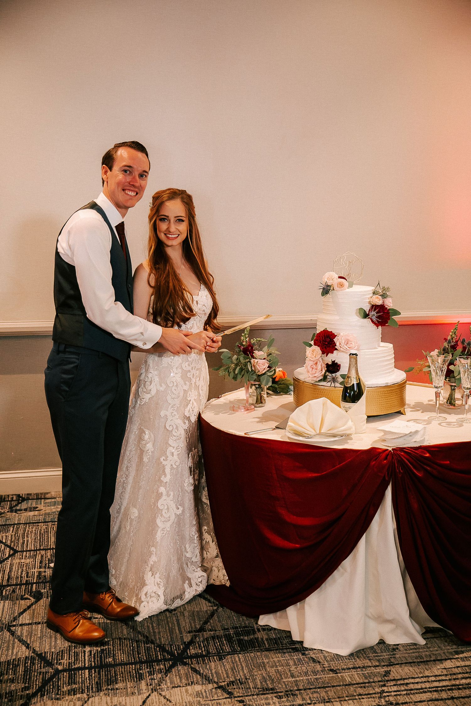 Bride and groom smiling while cutting their white wedding cake at a reception with a burgundy velvet tablecloth.