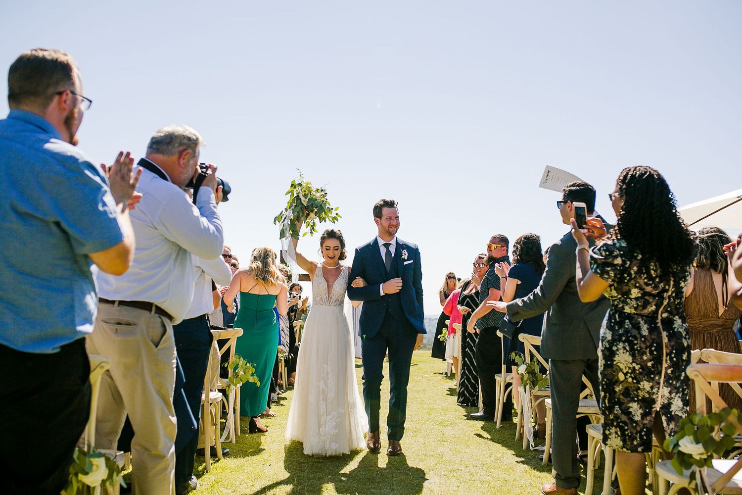 Joyful bride and groom walk down outdoor wedding aisle, guests clapping under clear blue sky.