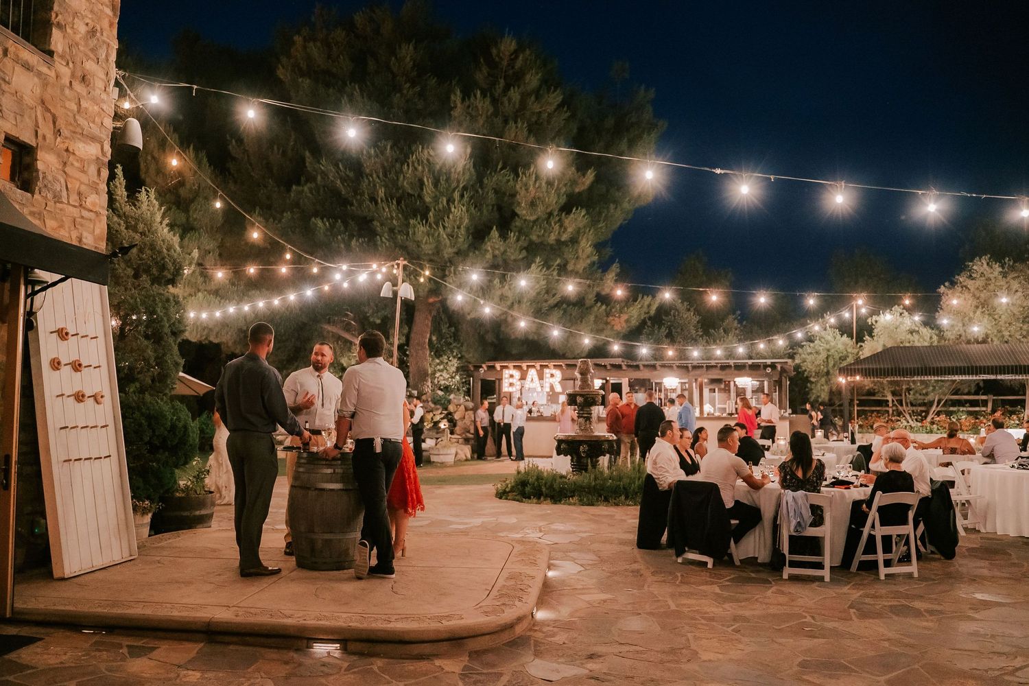 Outdoor wedding reception at night with string lights, guests dining at white-clothed tables, and a rustic bar area.