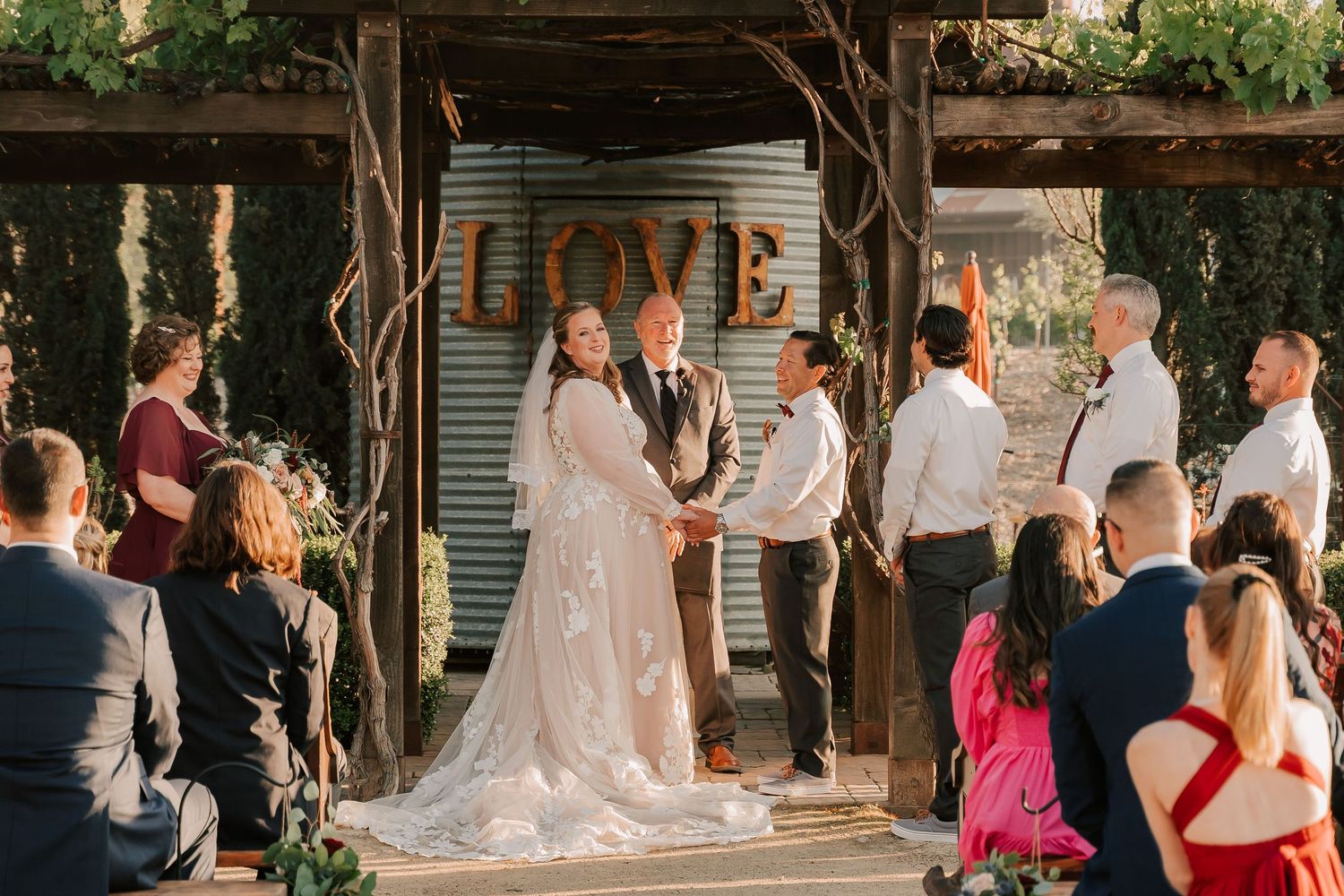 Outdoor rustic wedding ceremony with bride and groom at altar under LOVE sign, surrounded by guests and wedding party.