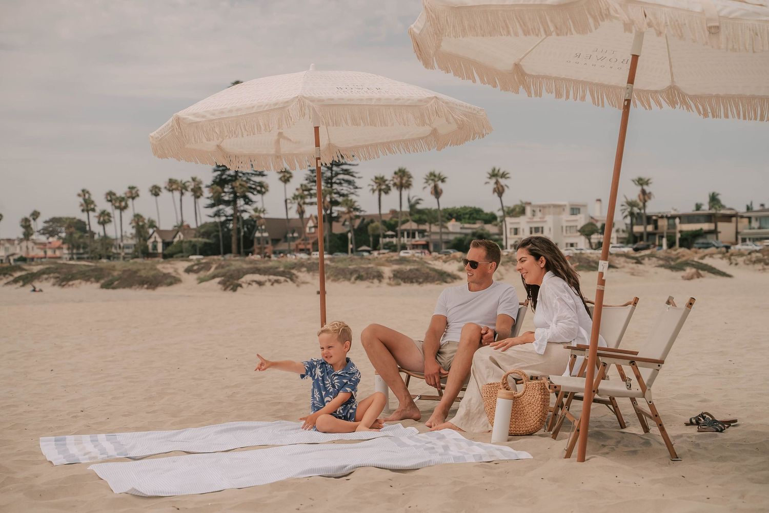 Family relaxing on a white blanket under fringed beach umbrellas on a sandy beach with palm trees.
