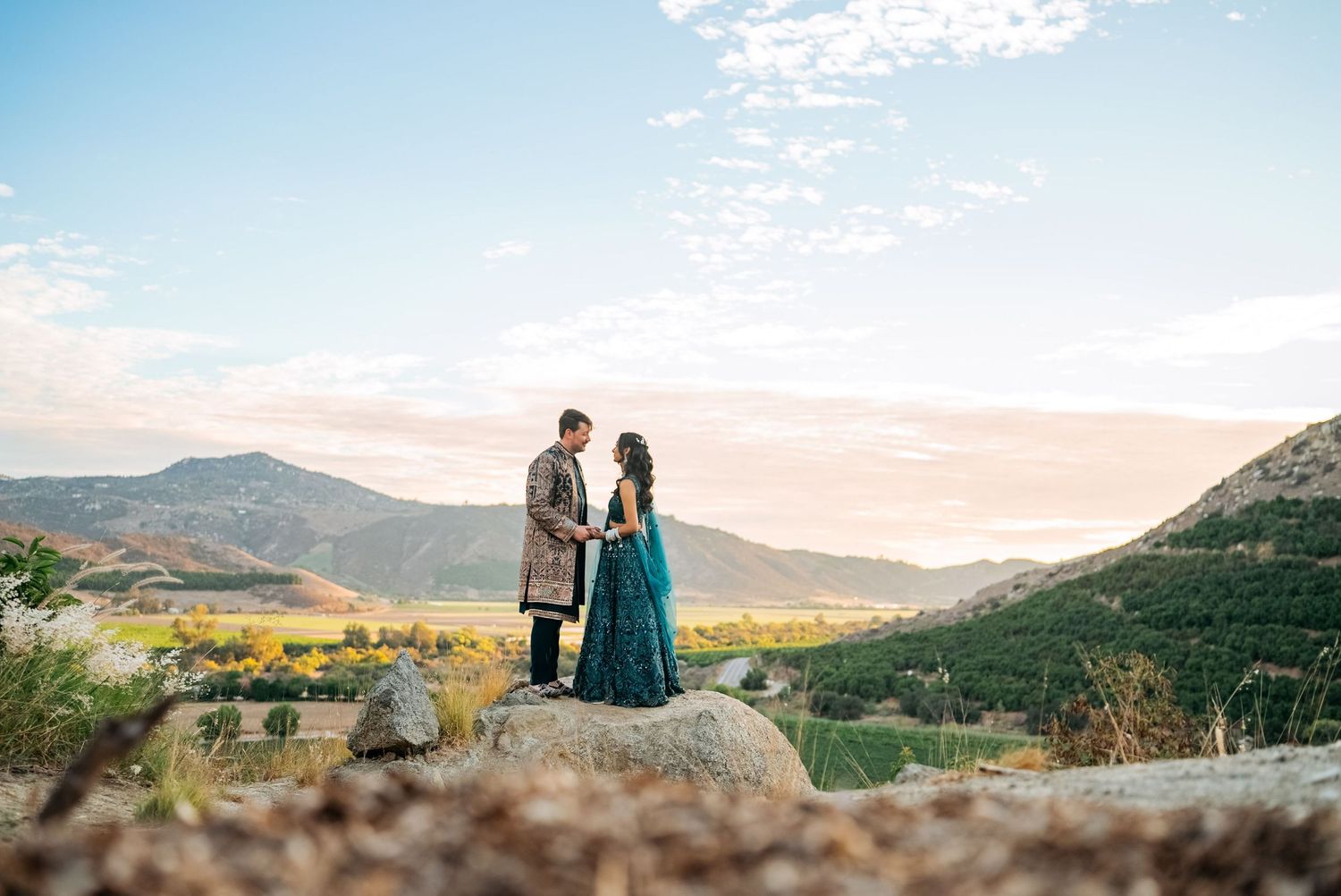 Couple in traditional Indian wedding attire standing on rocks with scenic mountain valley backdrop at sunset.