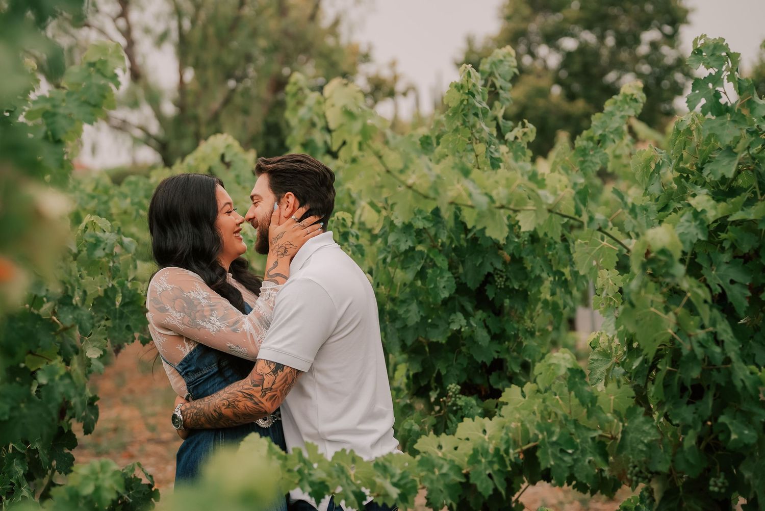 Couple sharing a romantic kiss among lush green grapevines in a vineyard, woman with tattoos embracing man.