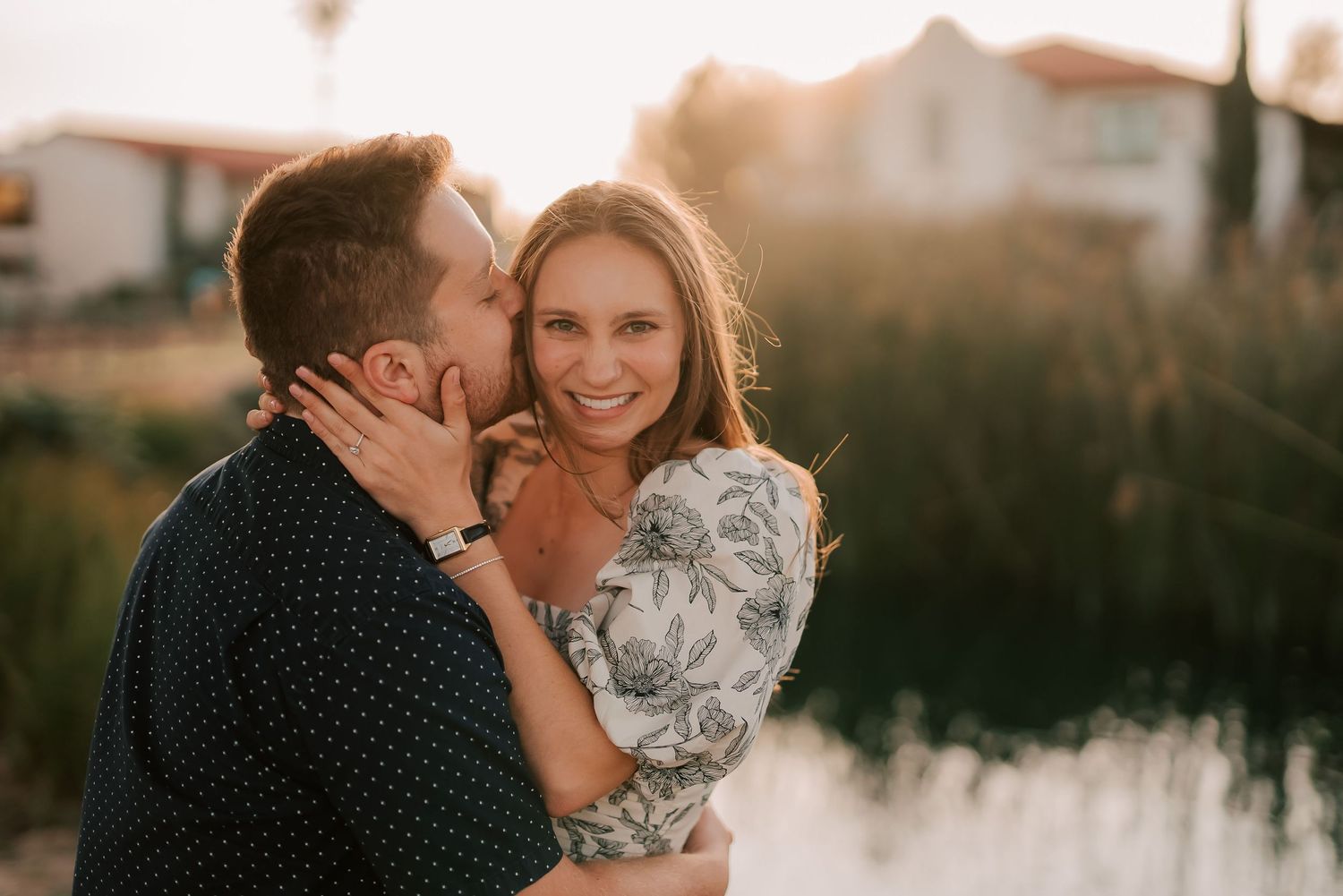 A couple shares a romantic sunset kiss outdoors near a lake, woman smiling warmly in a floral dress.