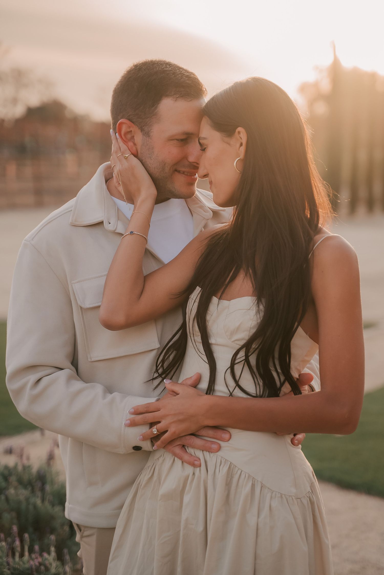 Smiling couple embracing forehead to forehead in a golden sunset glow, she in a cream dress and he in a light jacket.