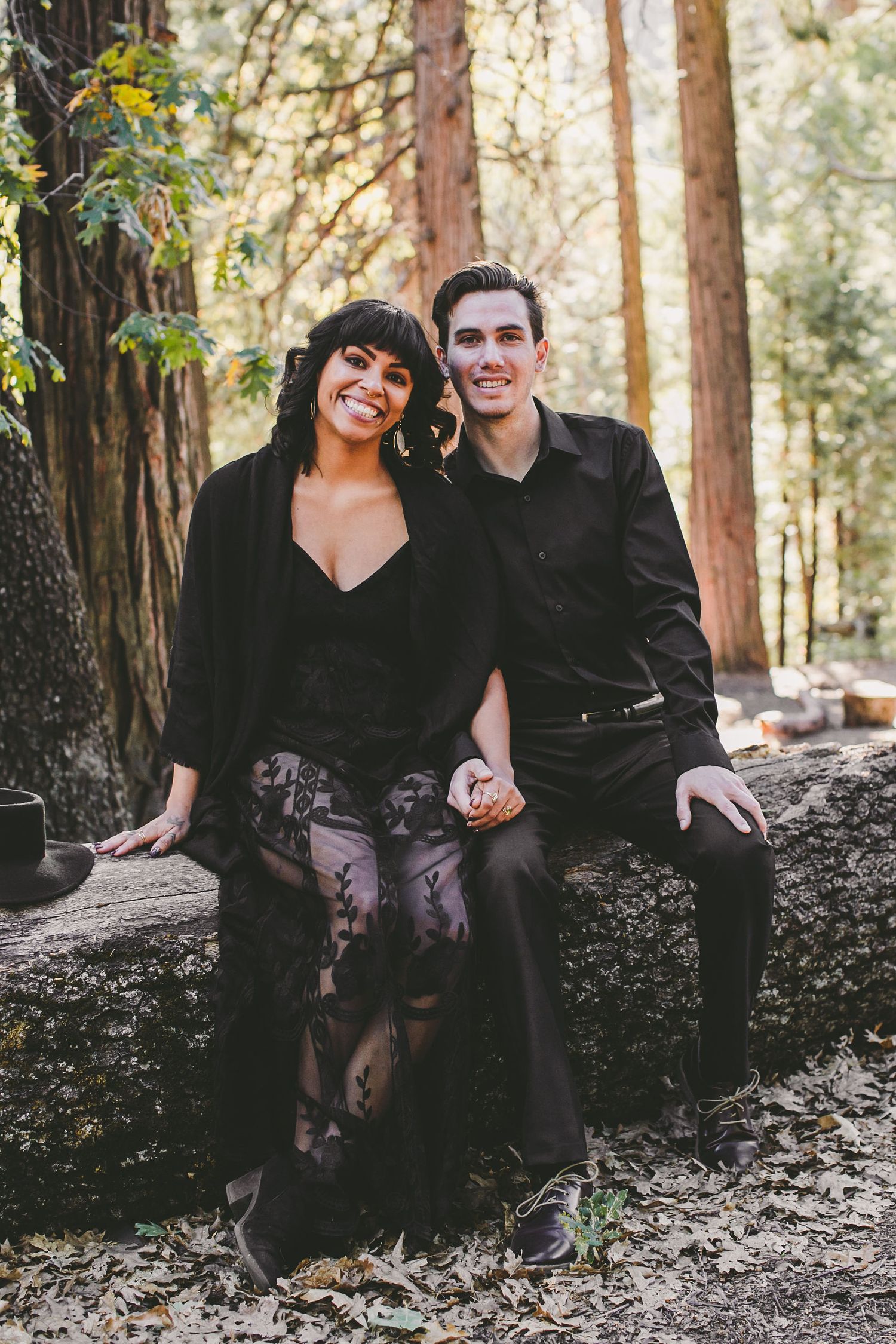 A smiling couple in matching black outfits sitting on a stone wall surrounded by tall trees in a forest setting.