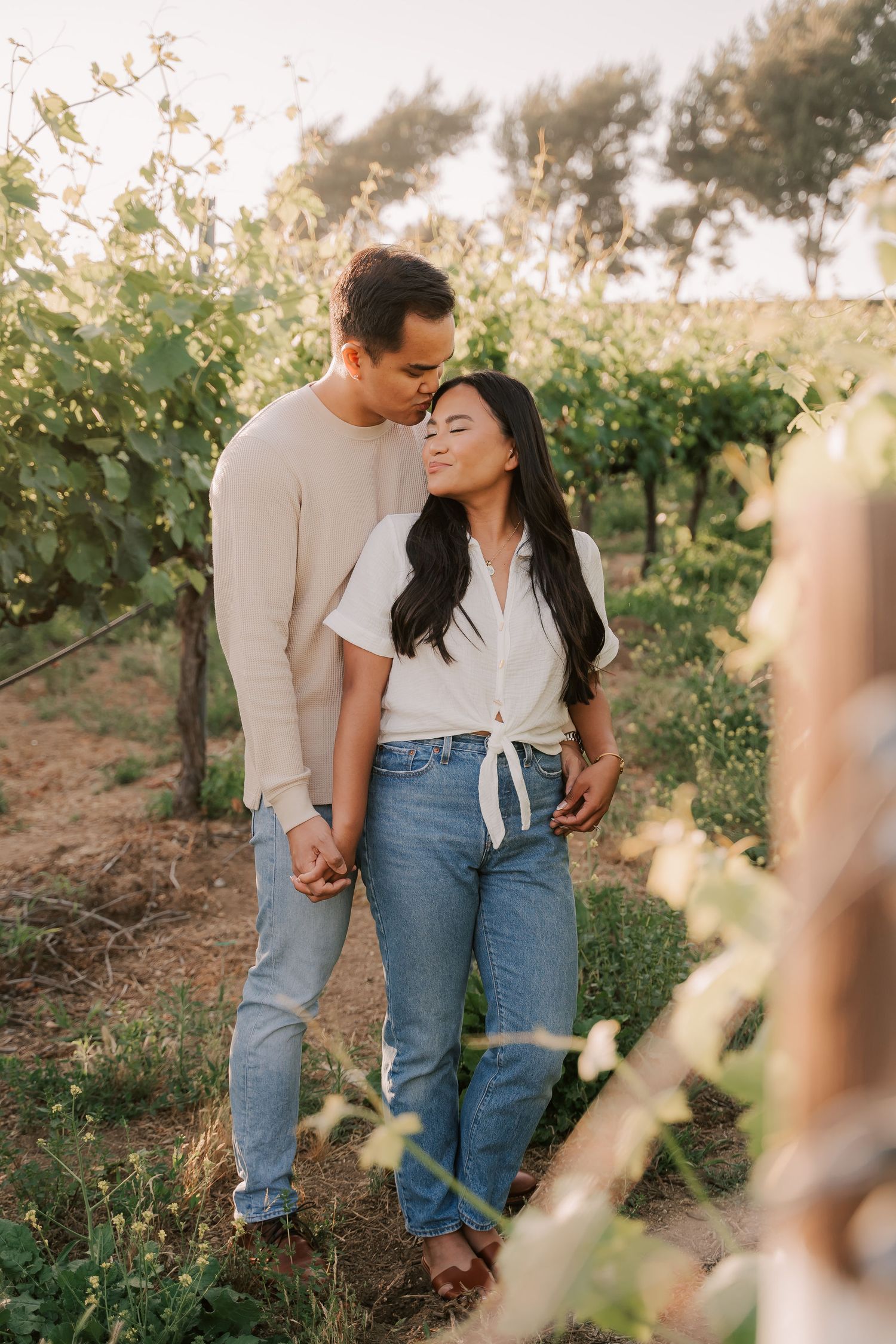 Couple holding hands and embracing in a lush vineyard at golden hour, wearing casual jeans and neutral tops.